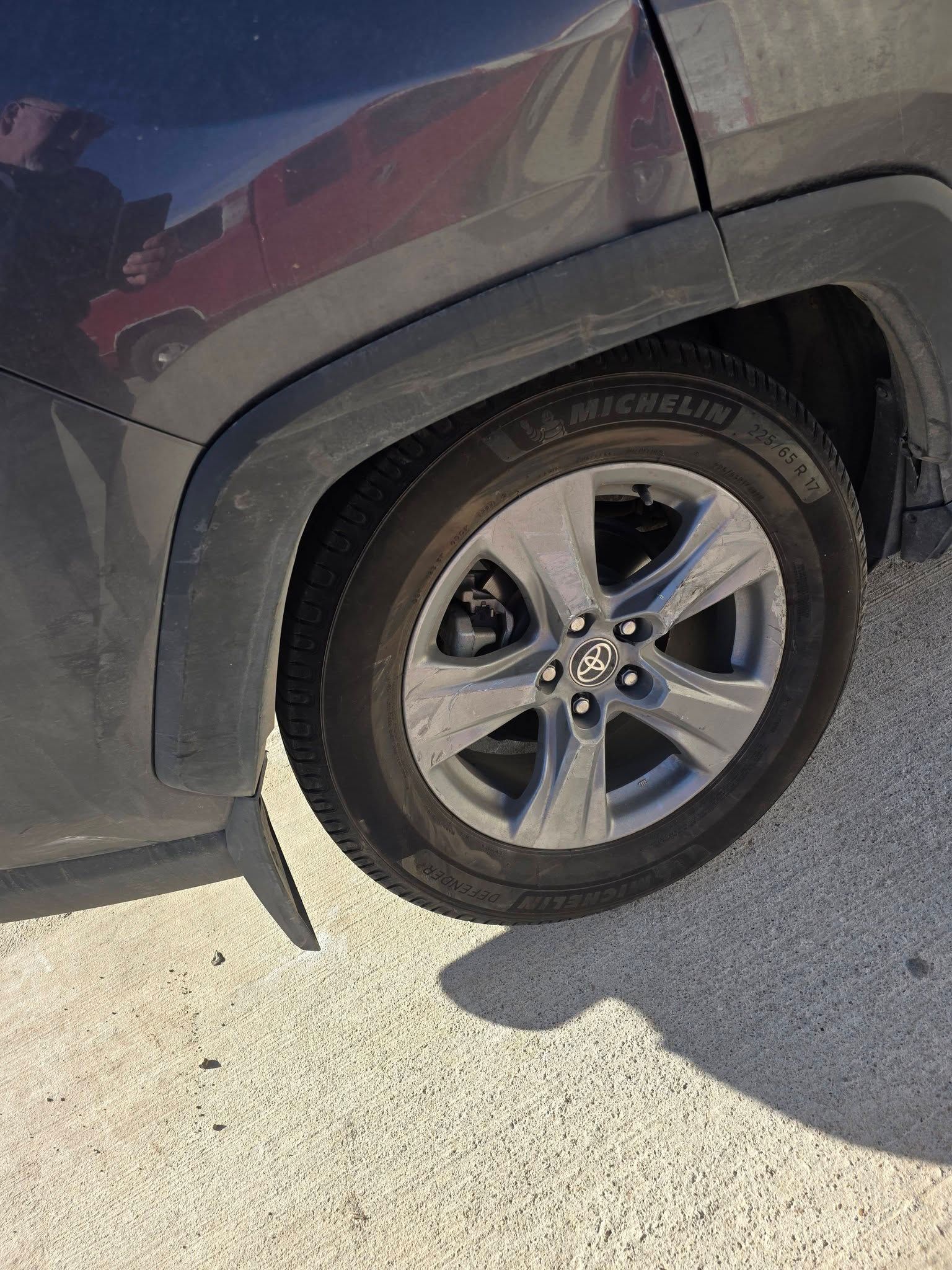 Close-up of a dark car’s rear wheel and tire on a bright pavement.