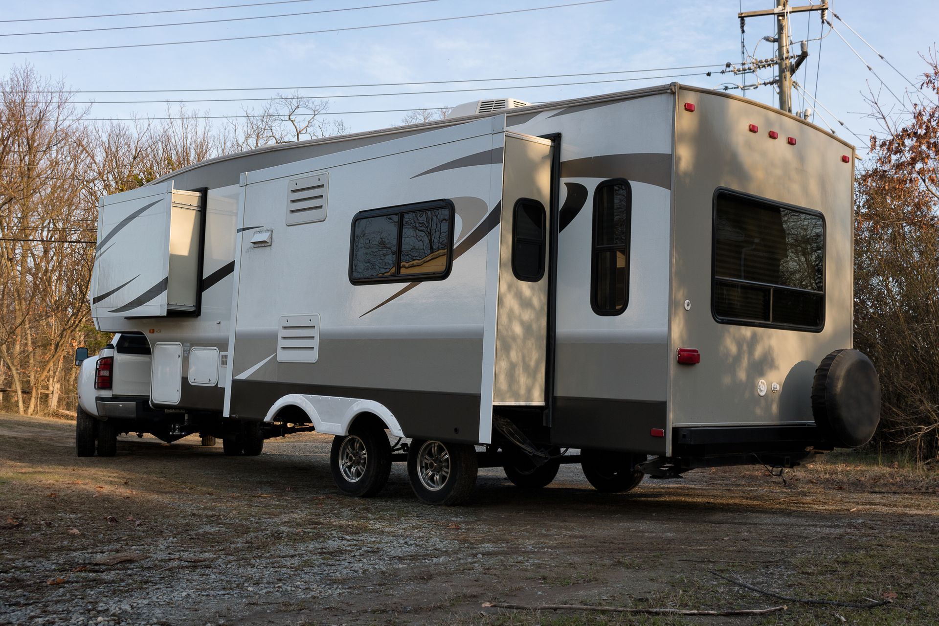White travel trailer parked on gravel outdoors beside trees, showing side and rear views.
