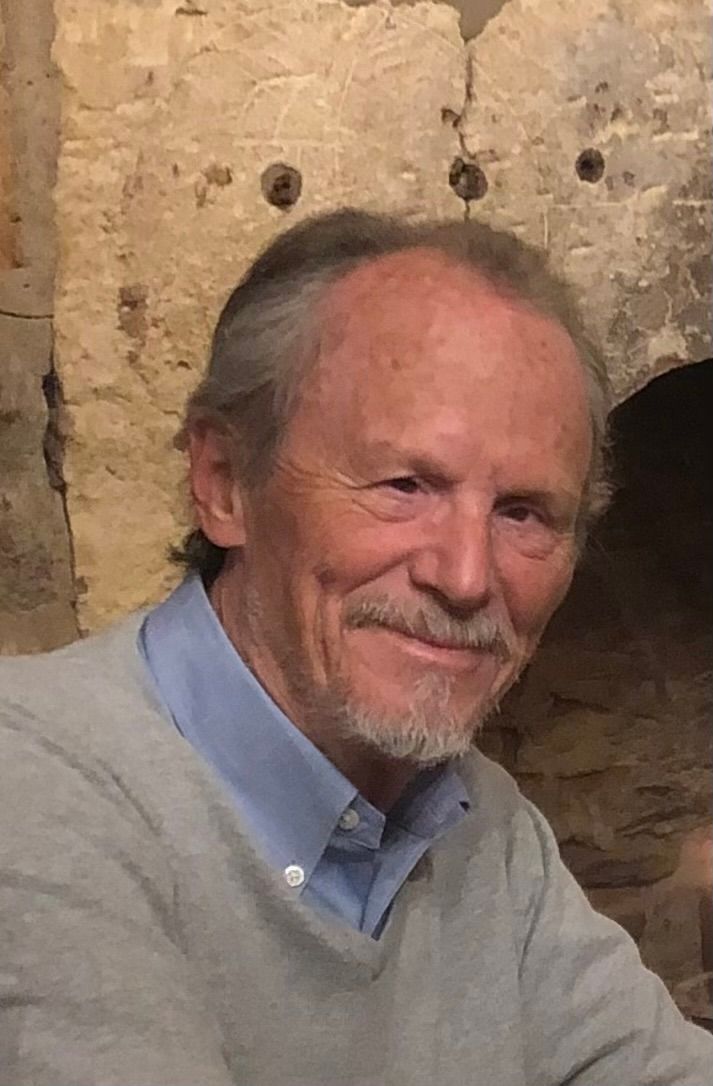 Smiling man in a light gray sweater and blue shirt against a stone wall