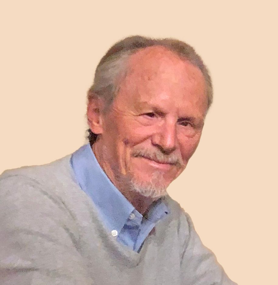 Smiling man in a light gray sweater and blue shirt against a stone wall