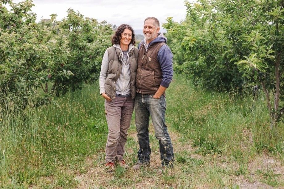Two people standing and smiling in a grassy field with trees behind them