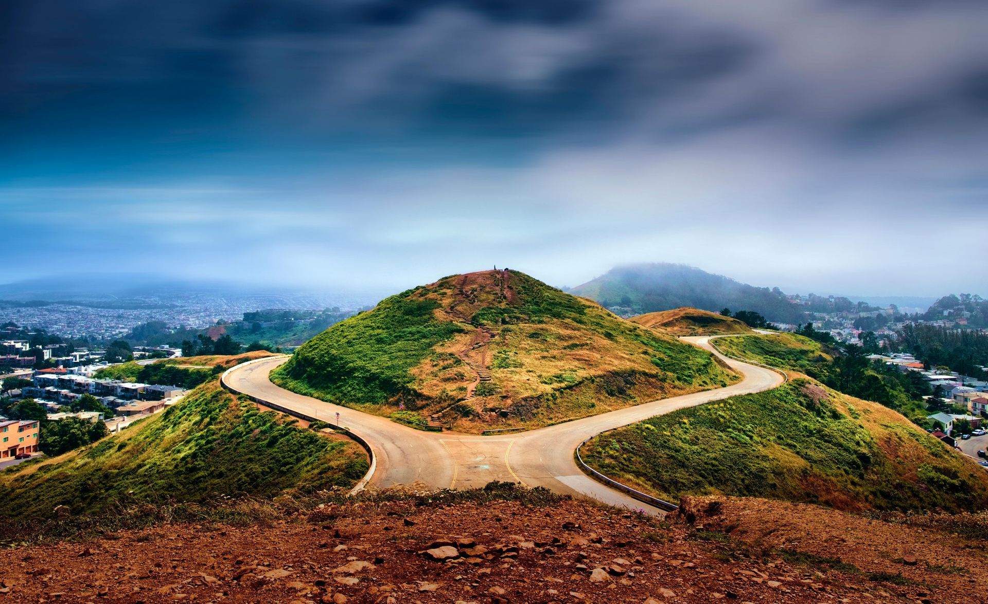 Winding dirt path up a green hill under a cloudy blue sky, overlooking a cityscape