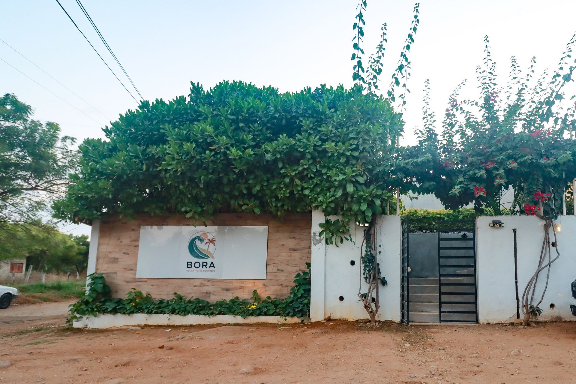 Entrada a la casa con portón blanco y letrero, enmarcada por árboles y plantas verdes en un camino de tierra.