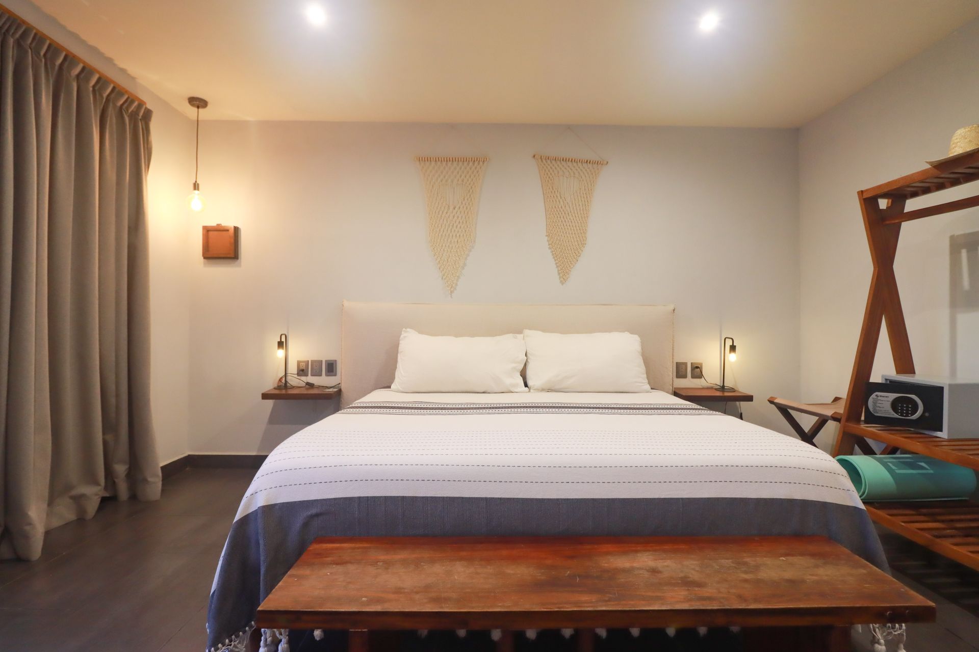 Dormitorio moderno con una cama bien hecha, lámparas de noche, cortinas y un banco de madera a los pies.