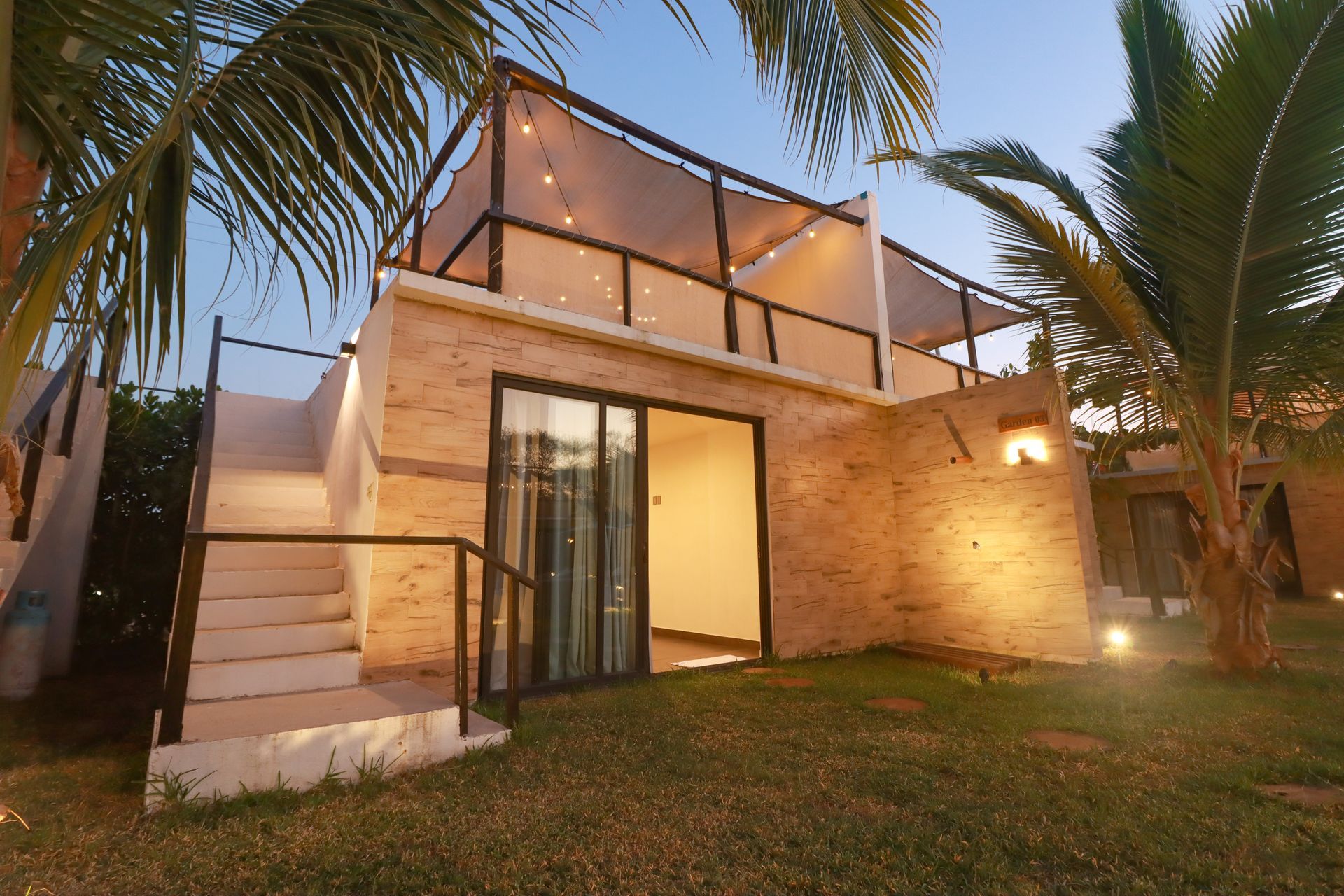 Villa moderna de dos plantas con iluminación cálida, balcón acristalado, muros de piedra y palmeras al atardecer.