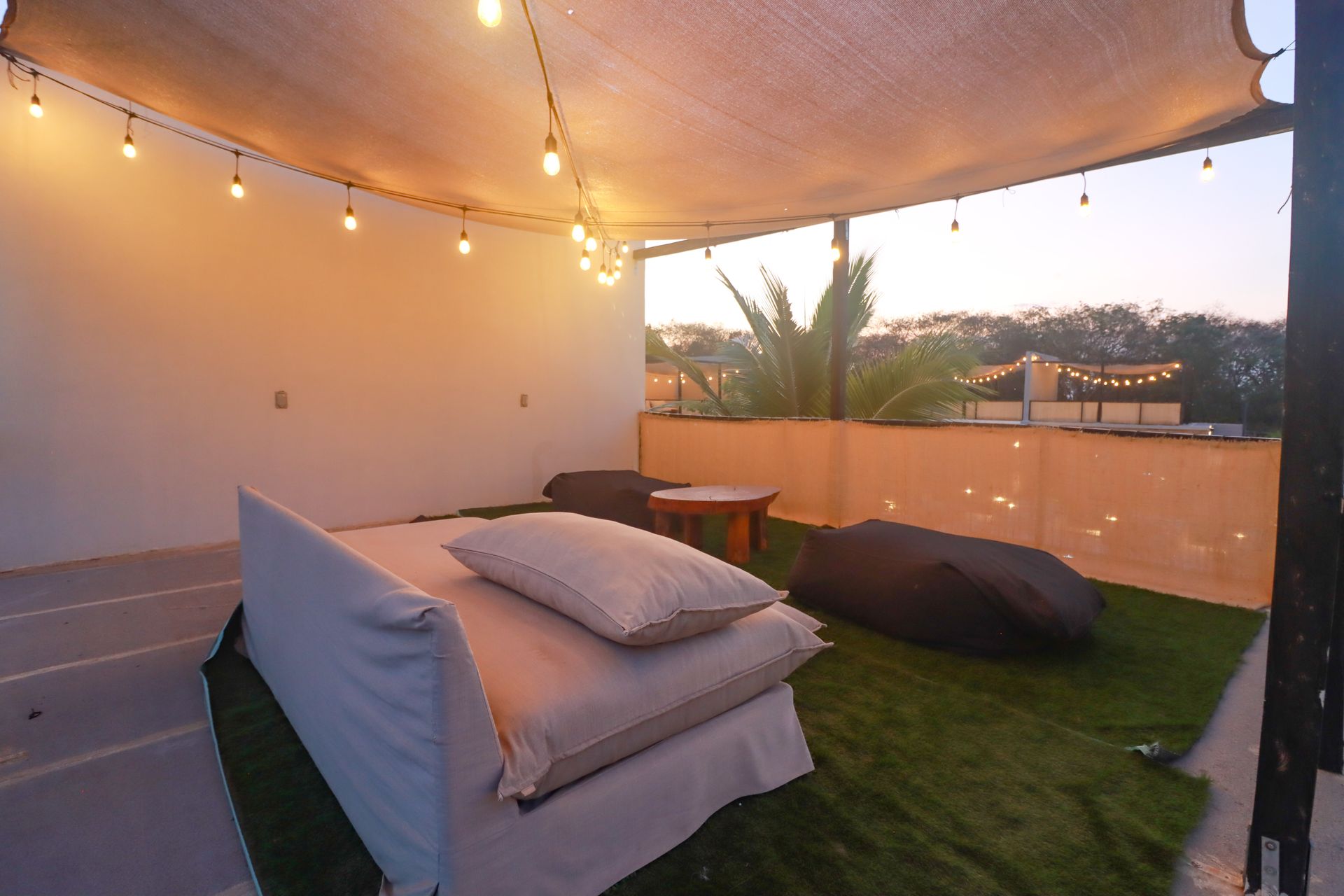 Terraza cubierta con luces de guirnalda, pufs y cojines sobre césped artificial al atardecer.