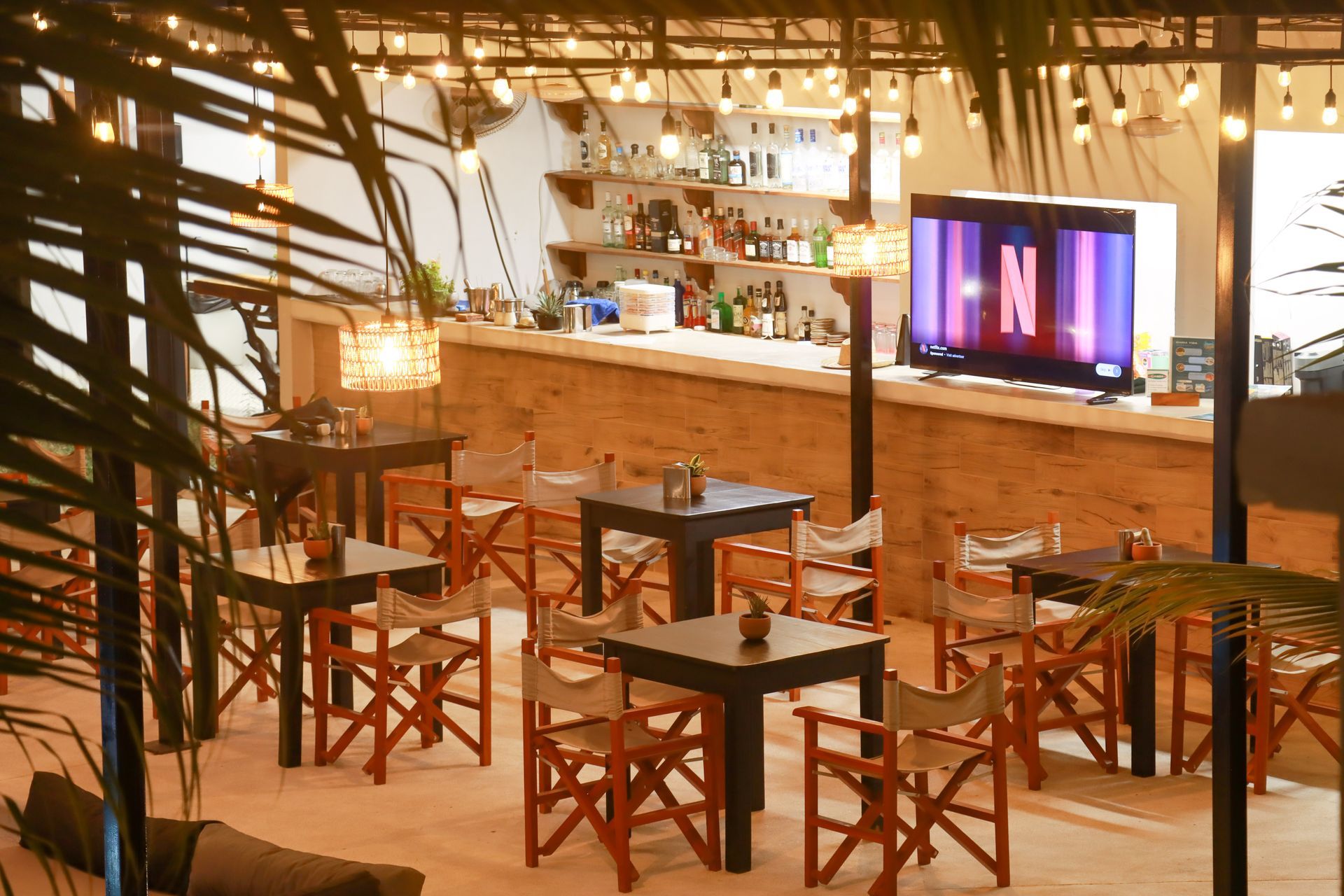 Interior de bar rústico con mesas y sillas de madera, iluminación cálida y un televisor en la pared del fondo.