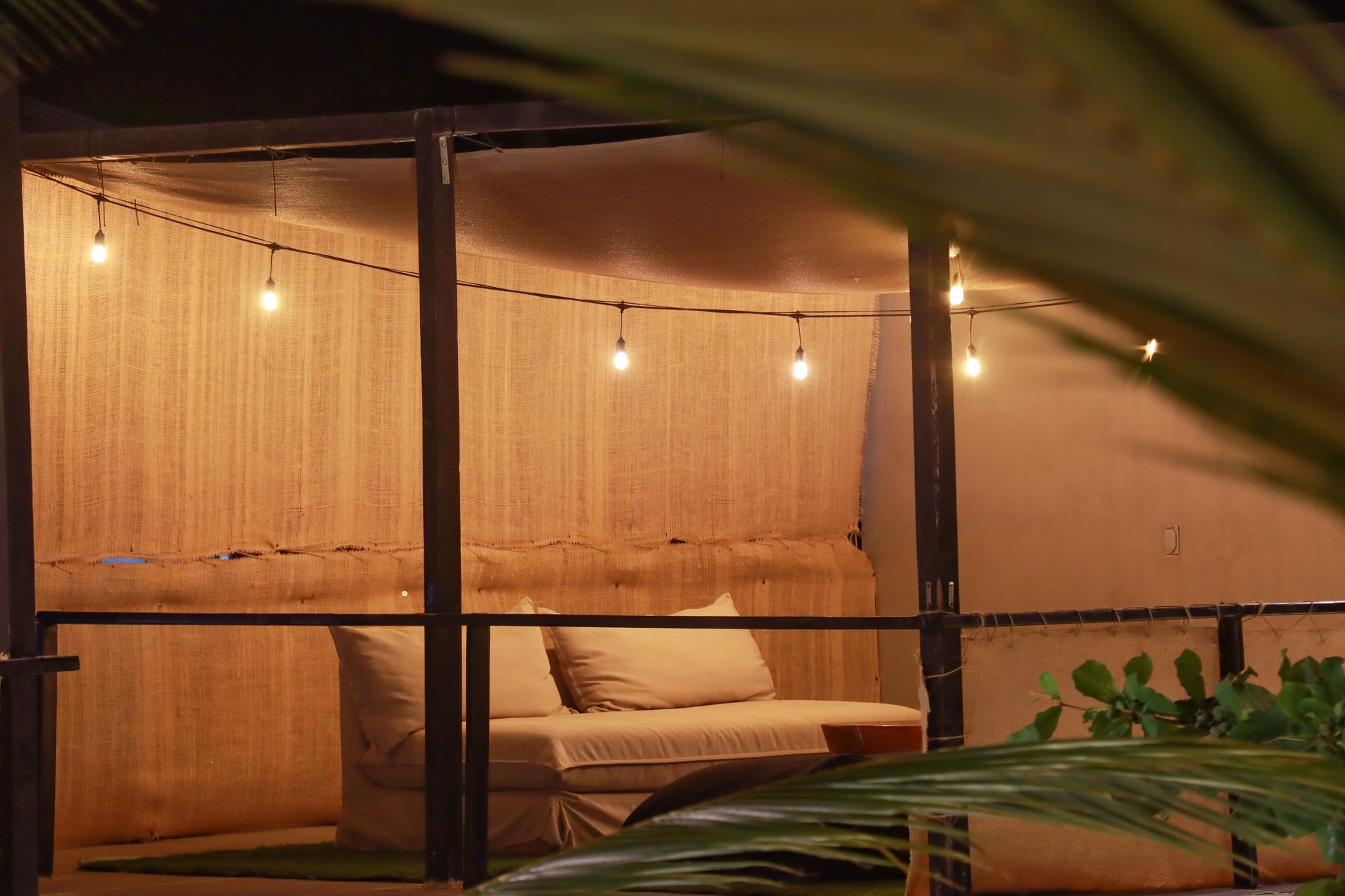 Acogedor salón al aire libre con sofás beige, guirnaldas de luces y plantas tropicales al atardecer.