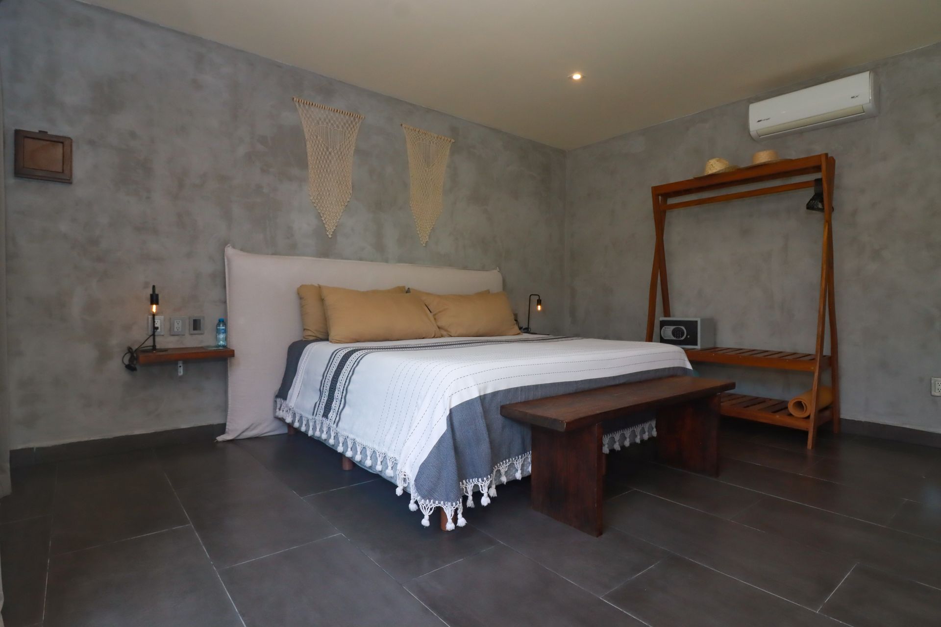 Dormitorio minimalista con cama blanca, banco de madera y perchero sobre suelo de baldosas oscuras.