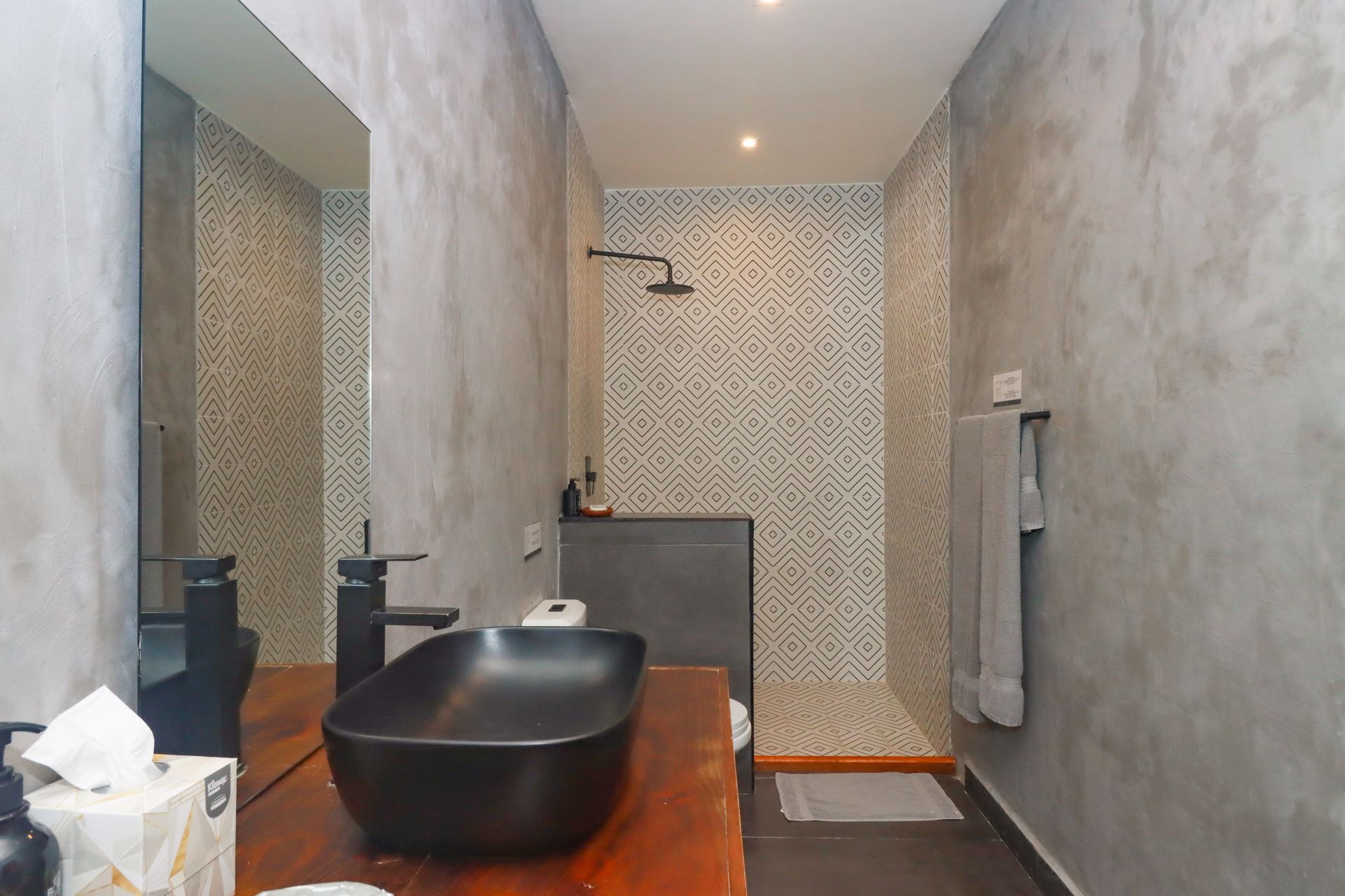 Baño moderno con lavabo de sobreponer negro, tocador de madera, ducha y pared de azulejos estampados.