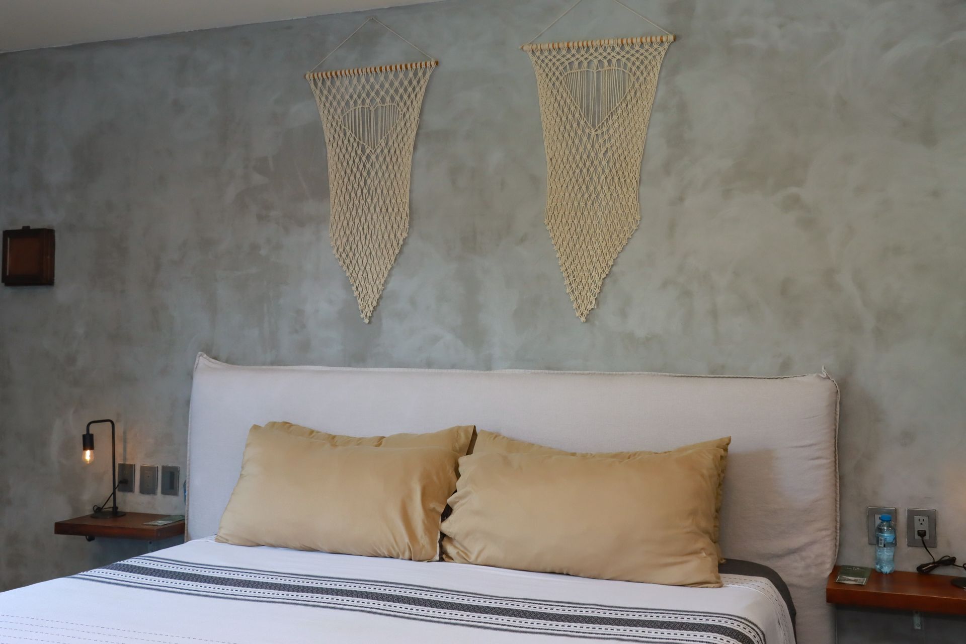 Dormitorio minimalista con una cama blanca, almohadas color canela y decoración de pared tejida sobre una pared gris.