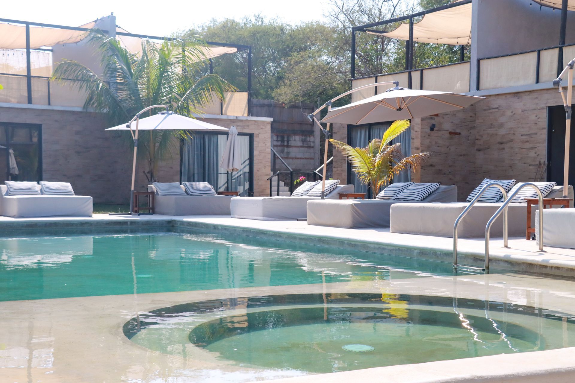 Piscina moderna tipo resort con tumbonas, sombrillas y cabañas bajo un cielo soleado.