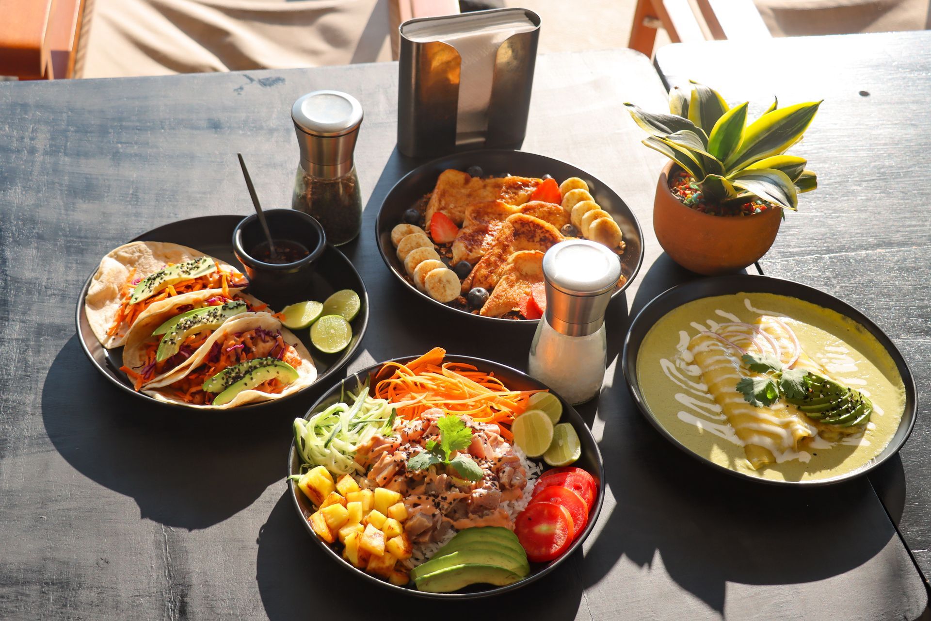 Mesa de exterior con cuatro platos coloridos, bebidas y una pequeña planta en maceta, bajo la luz del sol.