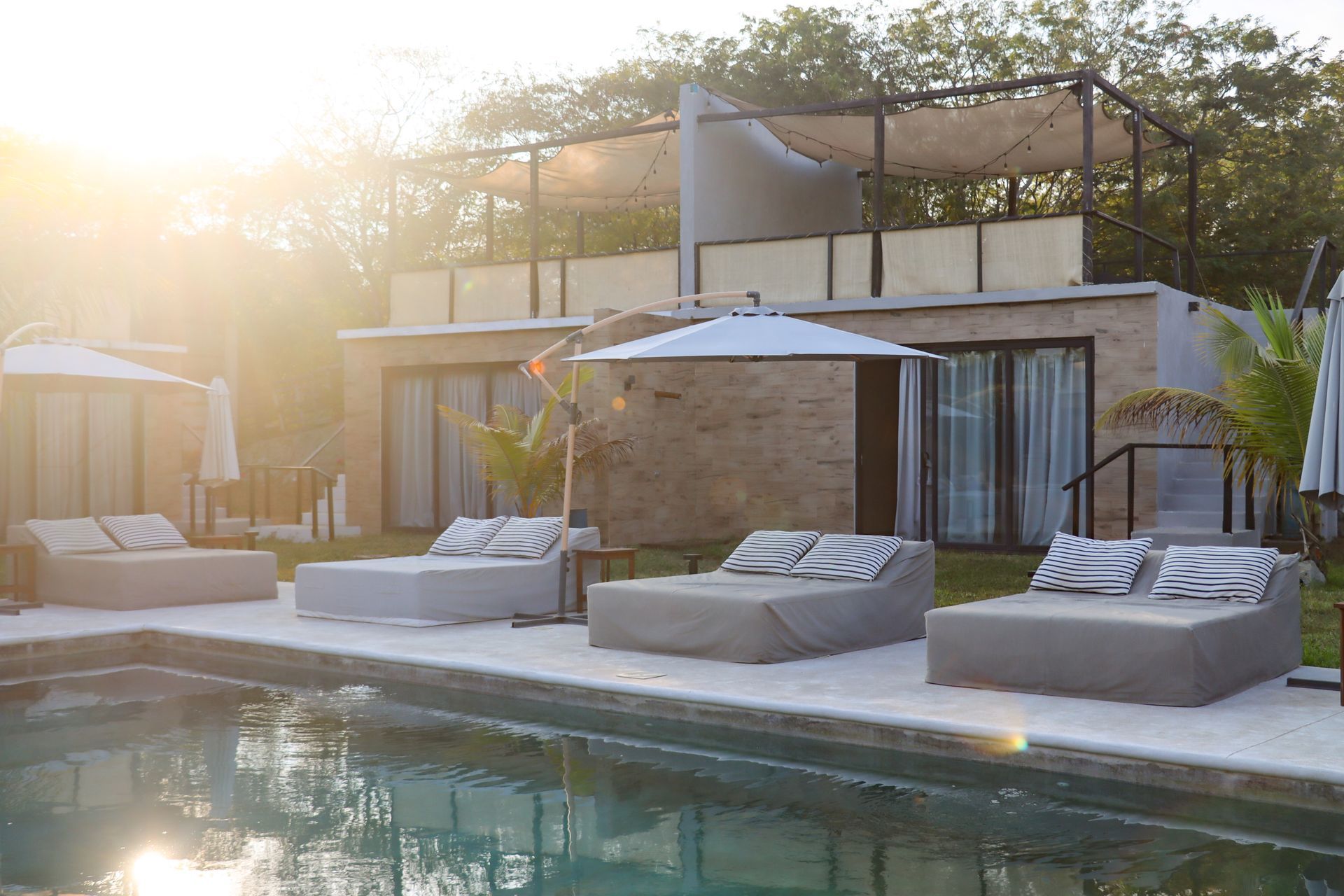 Moderno salón junto a la piscina con sombrillas blancas y tumbonas grises junto a una piscina iluminada por el sol.