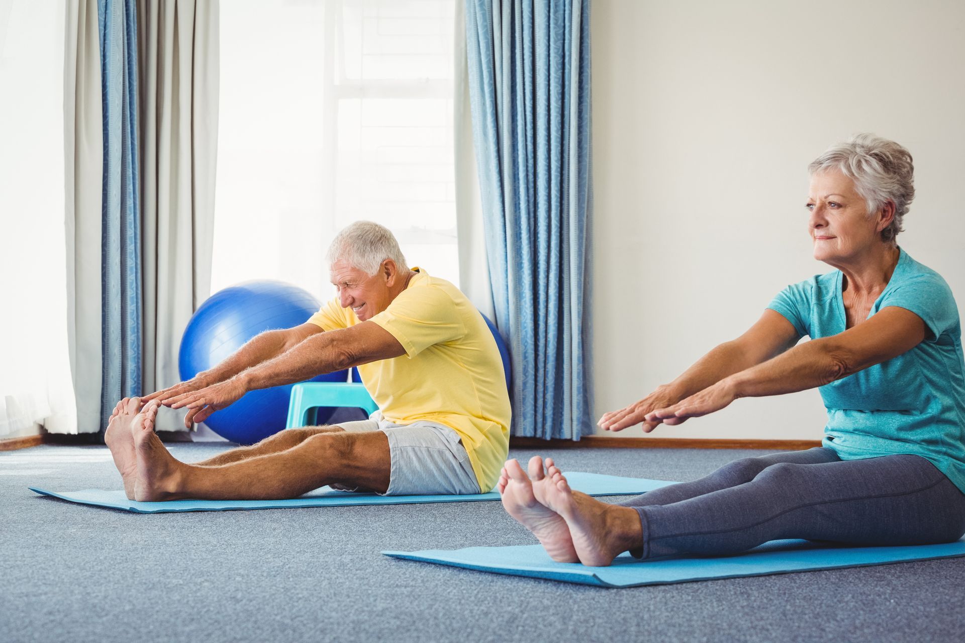 Two people stretching on mats in a bright exercise room, reaching toward their feet. Two people stretching on mats in a bright exercise room, reaching toward their feet.