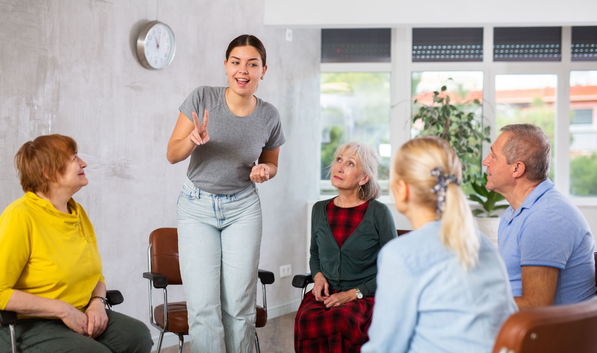 Woman speaking to a seated group in a bright room, with others listening attentively. Woman speaking to a seated group in a bright room, with others listening attentively.