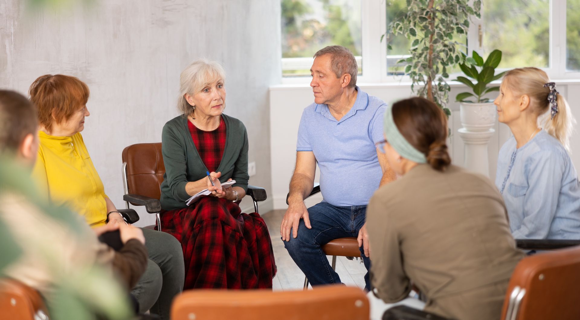 Small group seated in a circle, talking in a bright living room with plants and large windows. Small group seated in a circle, talking in a bright living room with plants and large windows.