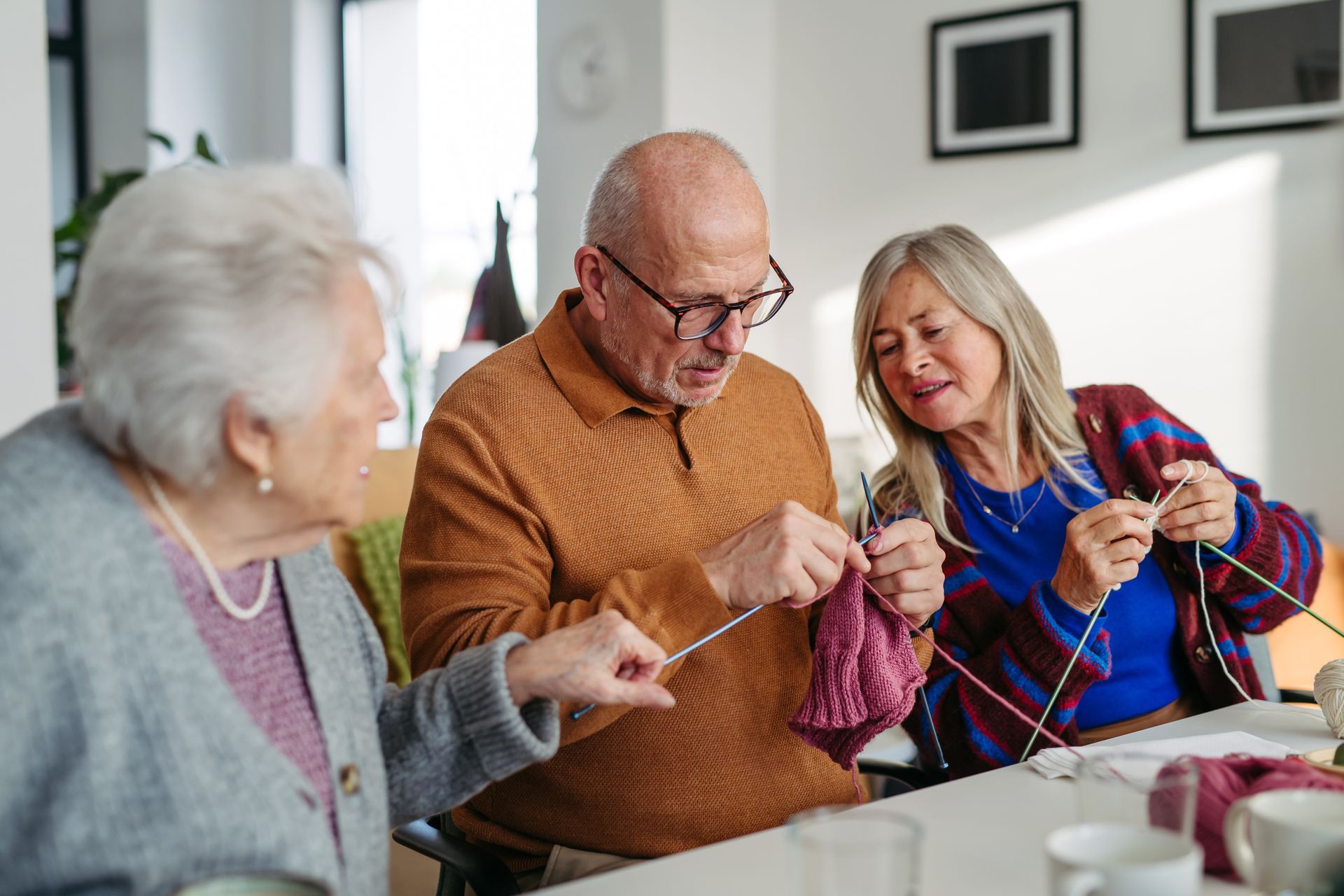 Three people knitting together at a table, smiling in a bright room. Three people knitting together at a table, smiling in a bright room.