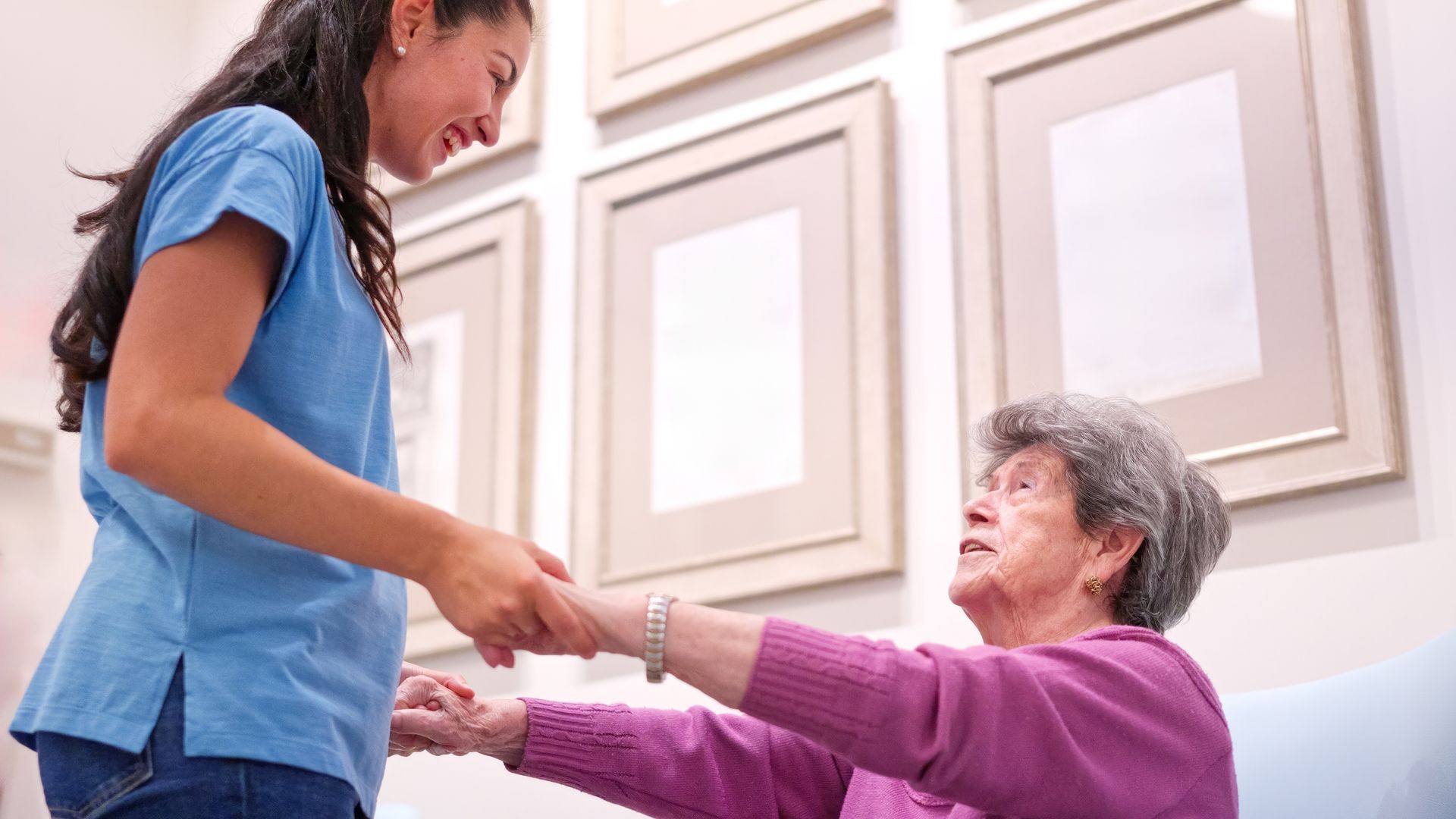 Young woman helping an elderly person stand in a bright room, holding hands and smiling Young woman helping an elderly person stand in a bright room, holding hands and smiling
