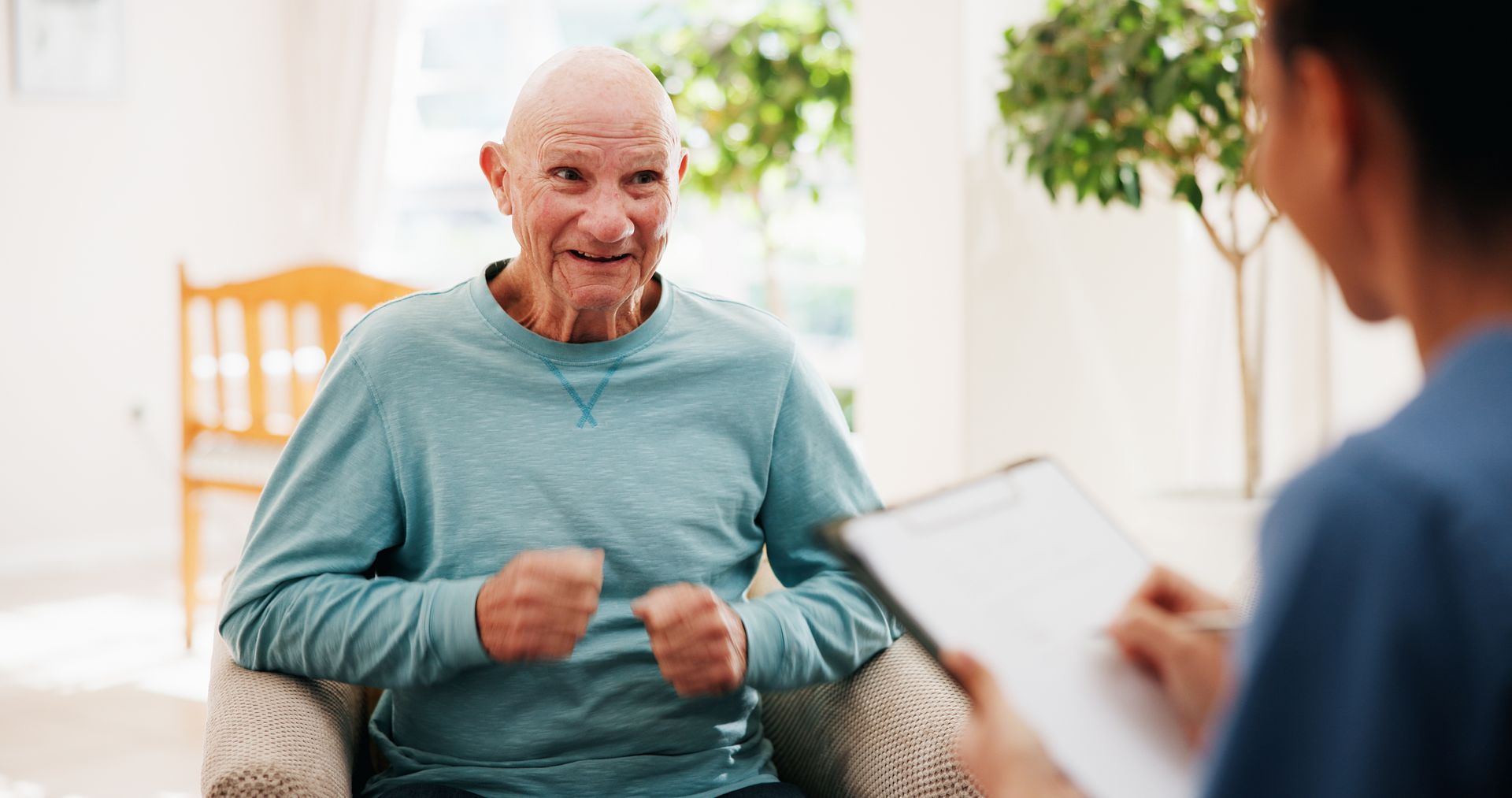 Man in teal shirt smiling and gesturing while talking with a clinician in a bright living room Man in teal shirt smiling and gesturing while talking with a clinician in a bright living room