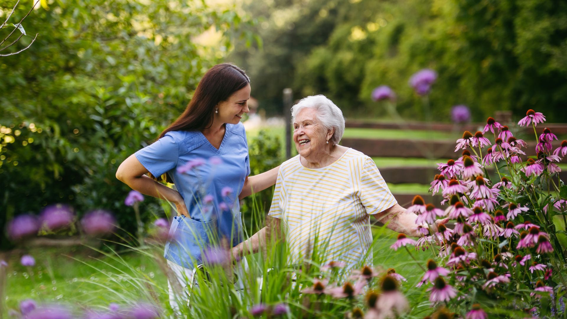 Two people standing in a lush garden, smiling among pink flowers and greenery.