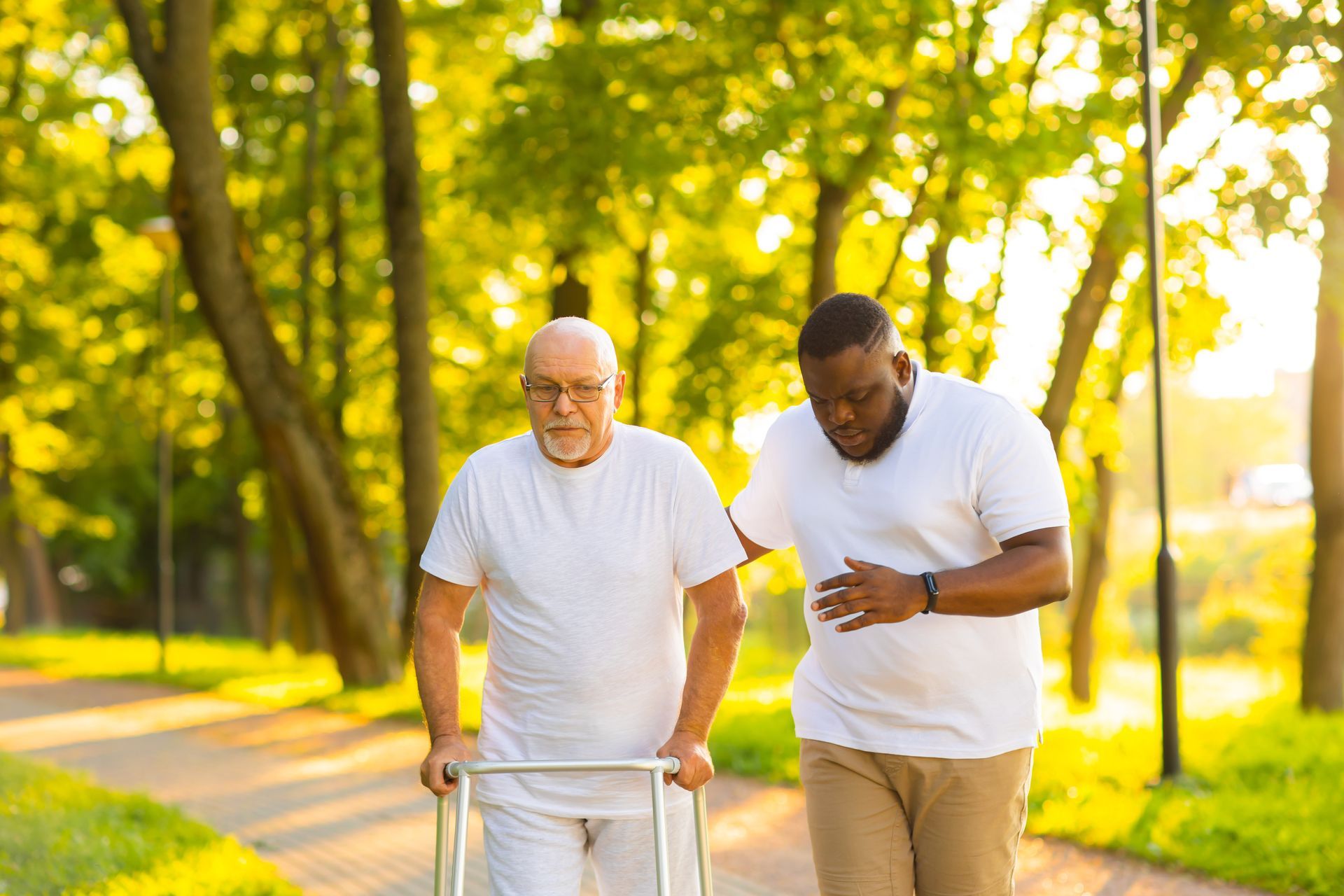Two men walking outdoors in a sunny park, one using a walker and the other supporting him Two men walking outdoors in a sunny park, one using a walker and the other supporting him