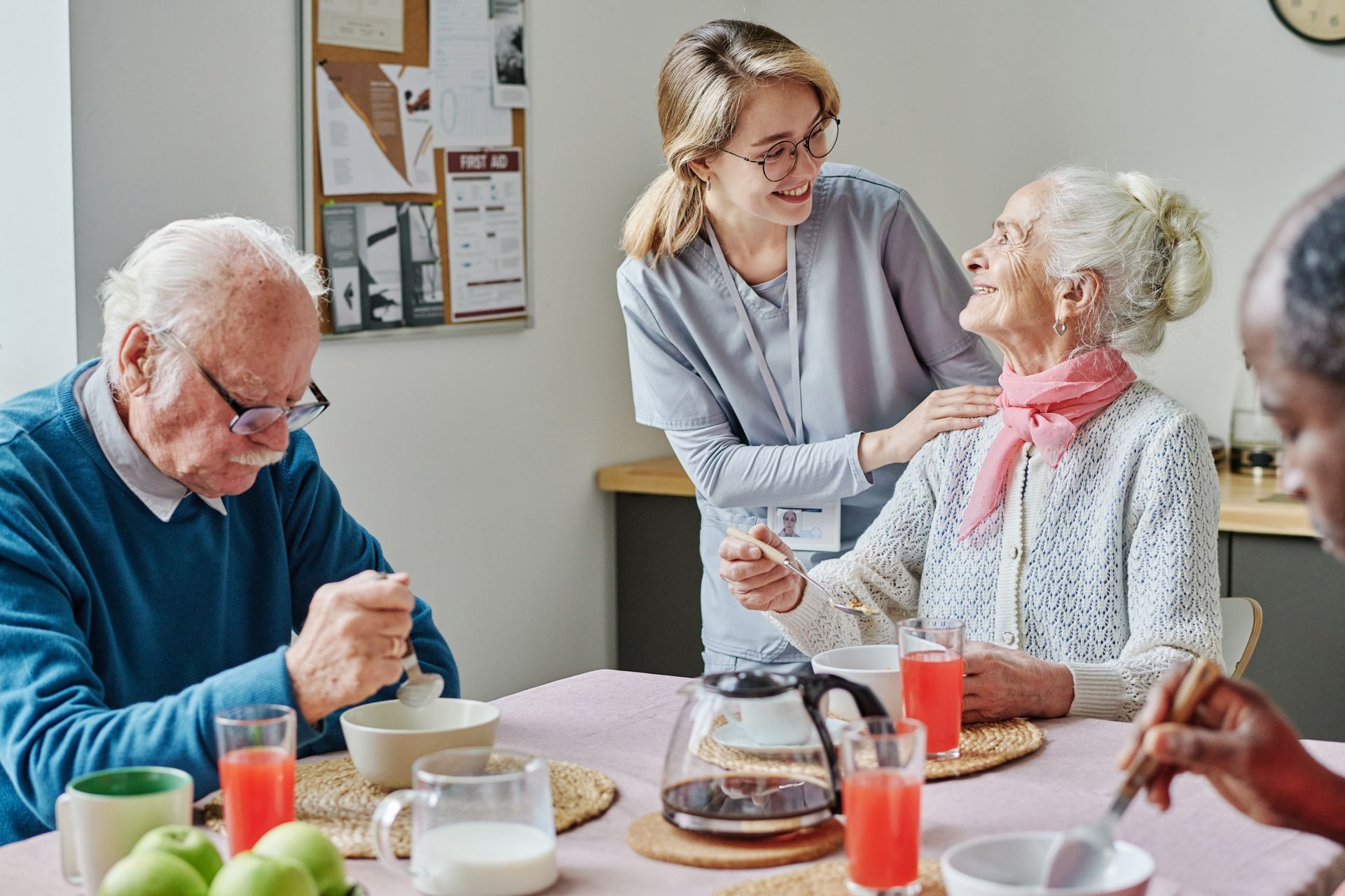 People sharing breakfast at a table, with a caregiver smiling beside an elderly woman in a bright scarf People sharing breakfast at a table, with a caregiver smiling beside an elderly woman in a bright scarf