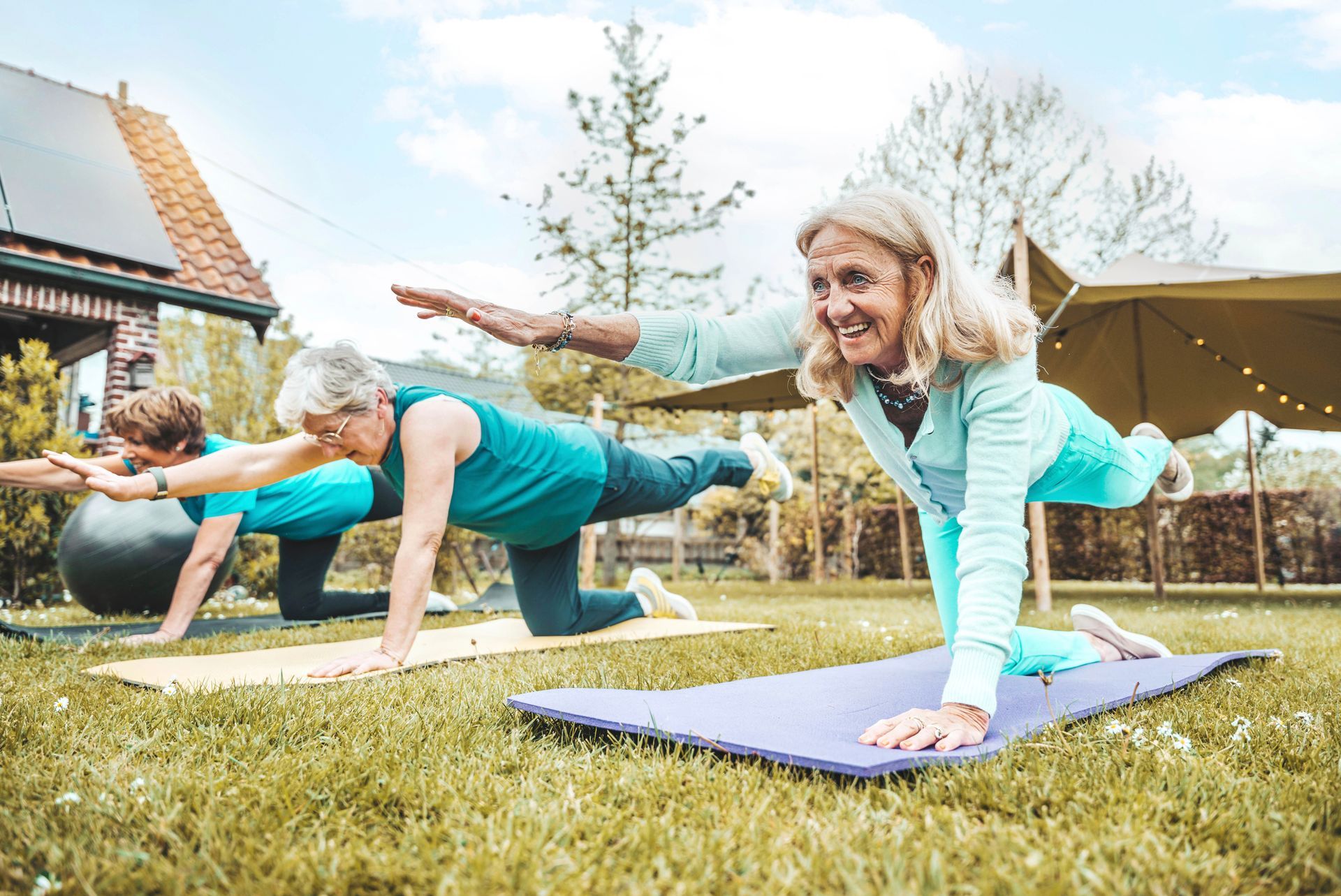 Two women doing yoga outdoors on mats in a grassy yard, with a house and canopy in the background Two women doing yoga outdoors on mats in a grassy yard, with a house and canopy in the background