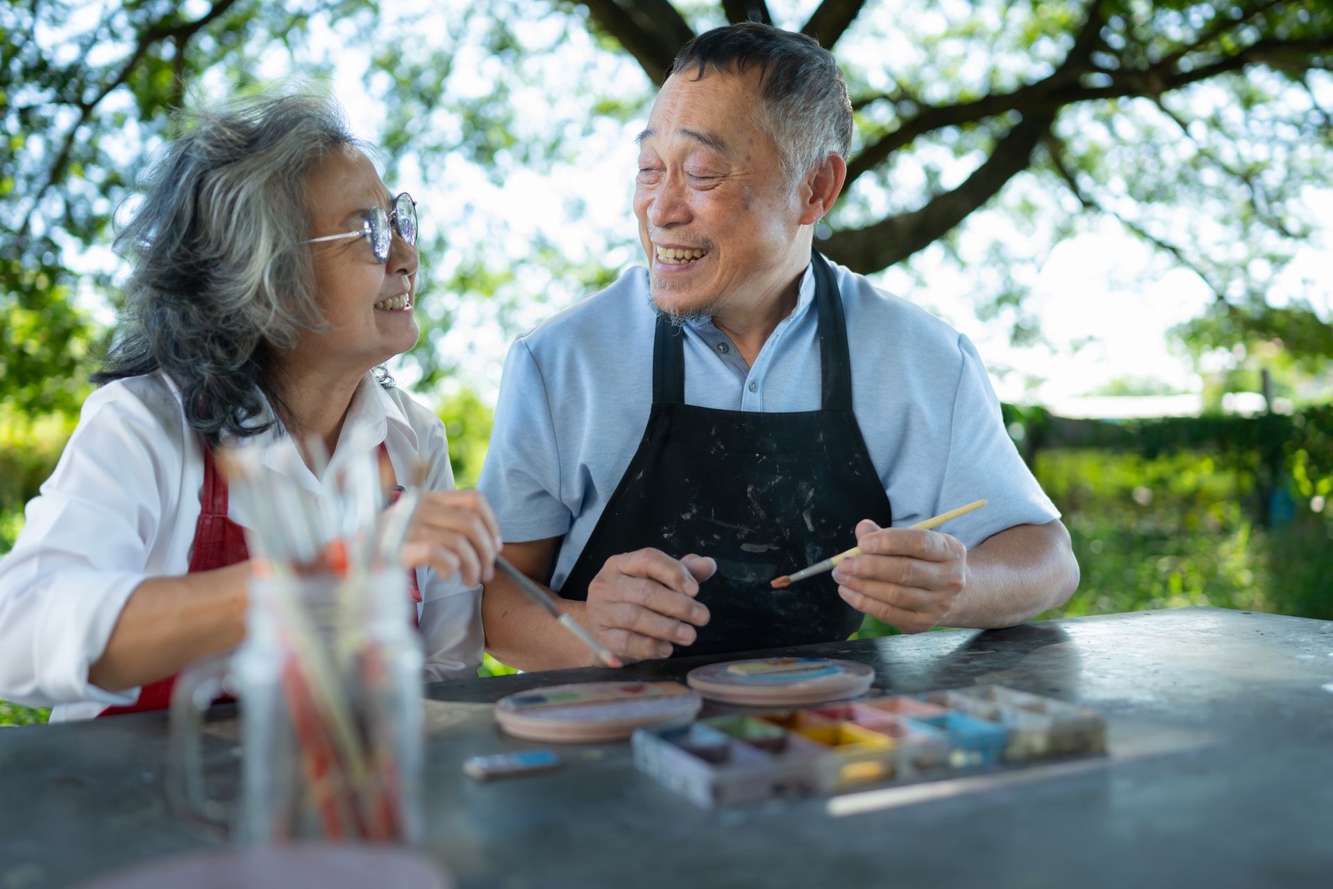 Two people smiling and painting together at a table outdoors under trees.