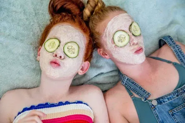 Two girls are laying on a bed with cucumber slices on their eyes.
