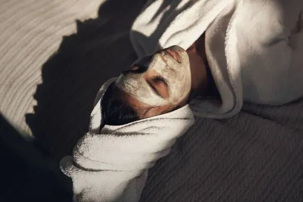 A woman is laying on a bed with a clay mask on her face.