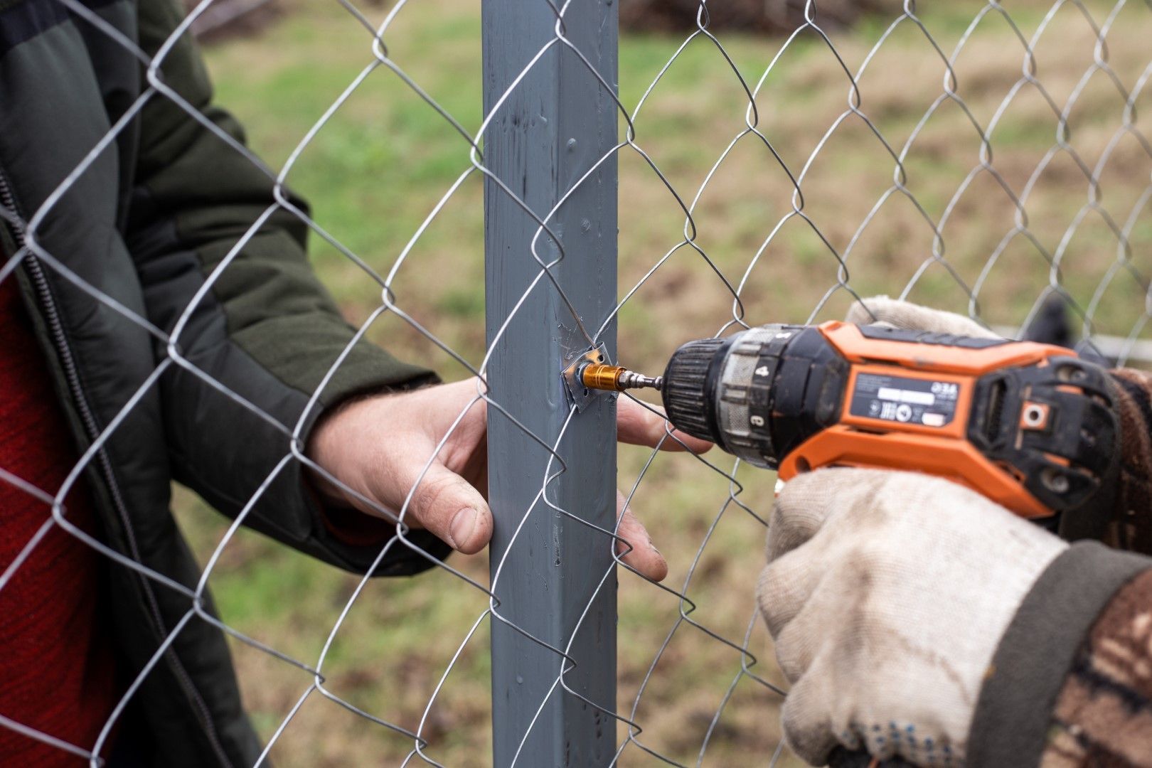Two people installing chain-link fence using a power drill on a gray post outdoors.