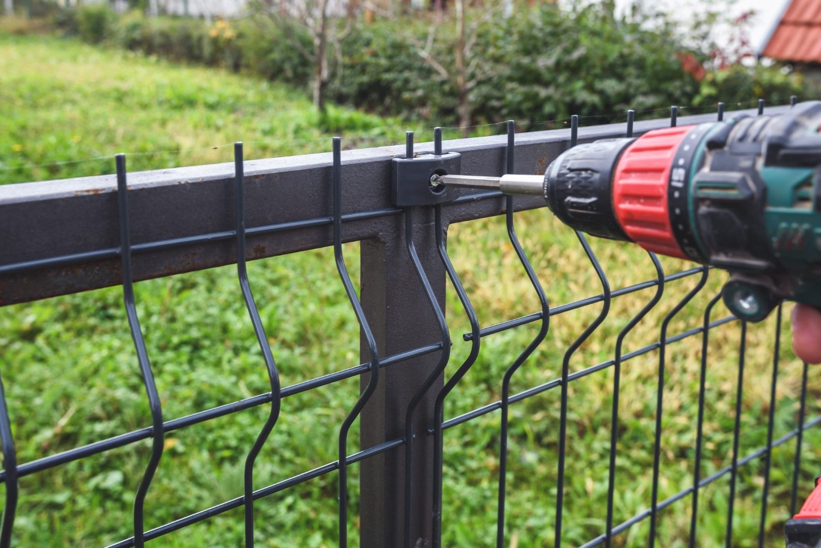 Person using a power drill to attach a black metal fence panel to a post outdoors on a grassy lawn.