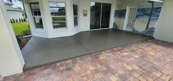 A newly poured concrete patio adjoins a brick patio and a white house with windows and a sliding glass door.