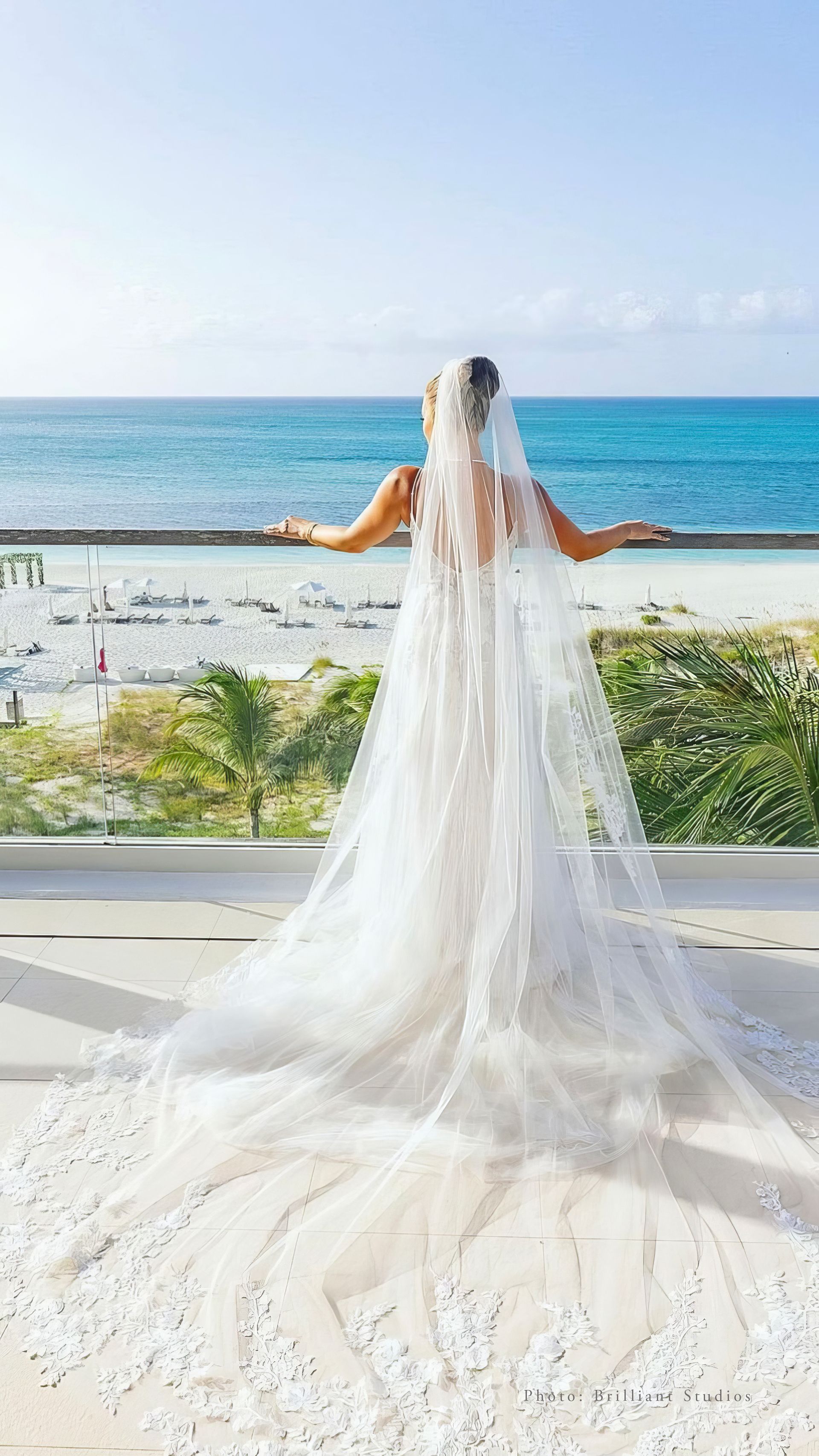 A bride in a wedding dress and veil is standing on a balcony overlooking the ocean.