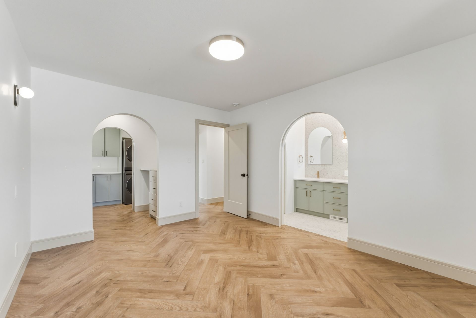 Empty white room with arched doorways, light wood herringbone floors, and a ceiling light.