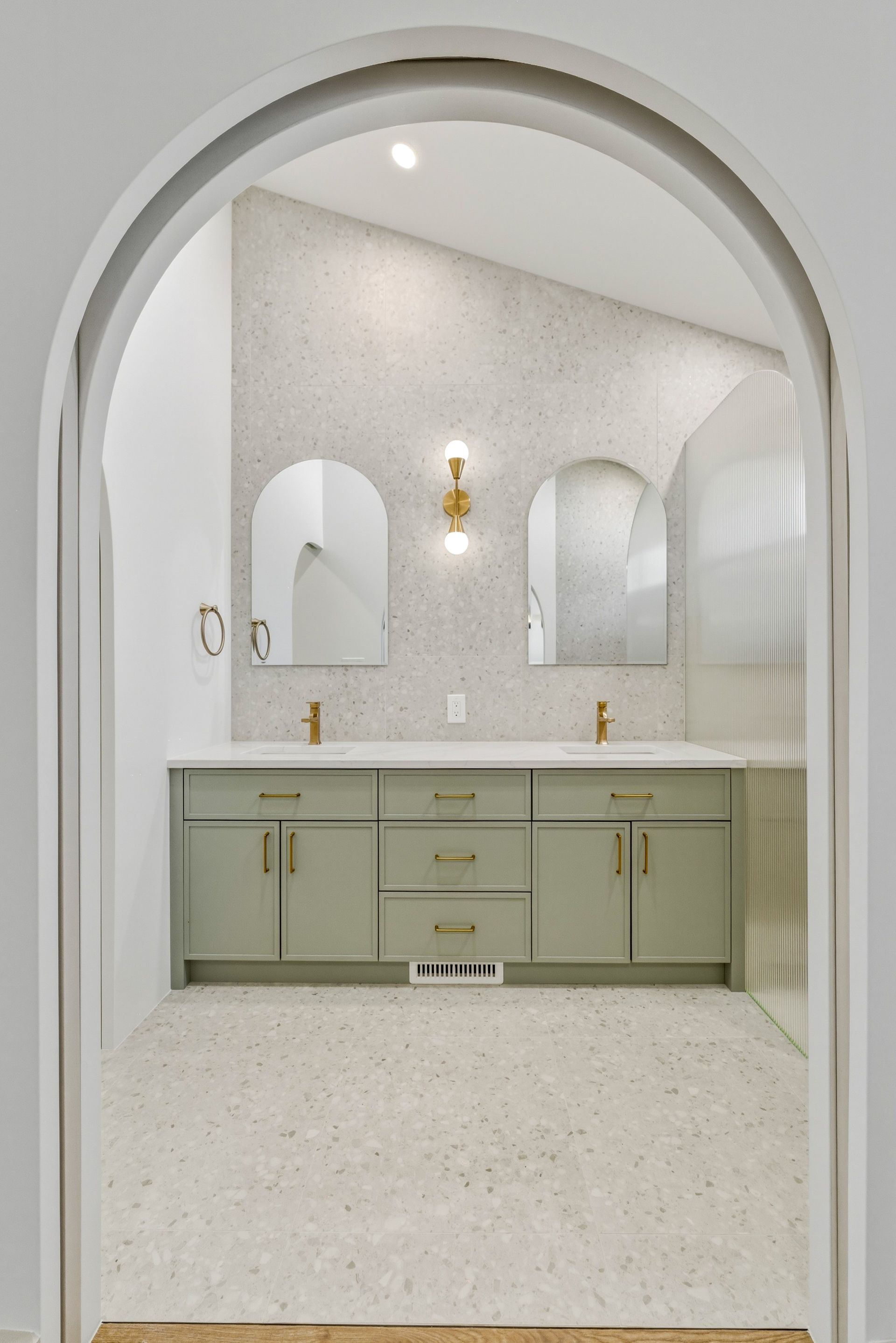 Arched bathroom vanity with pale green cabinets, gold fixtures, and mirrors in a bright white room
