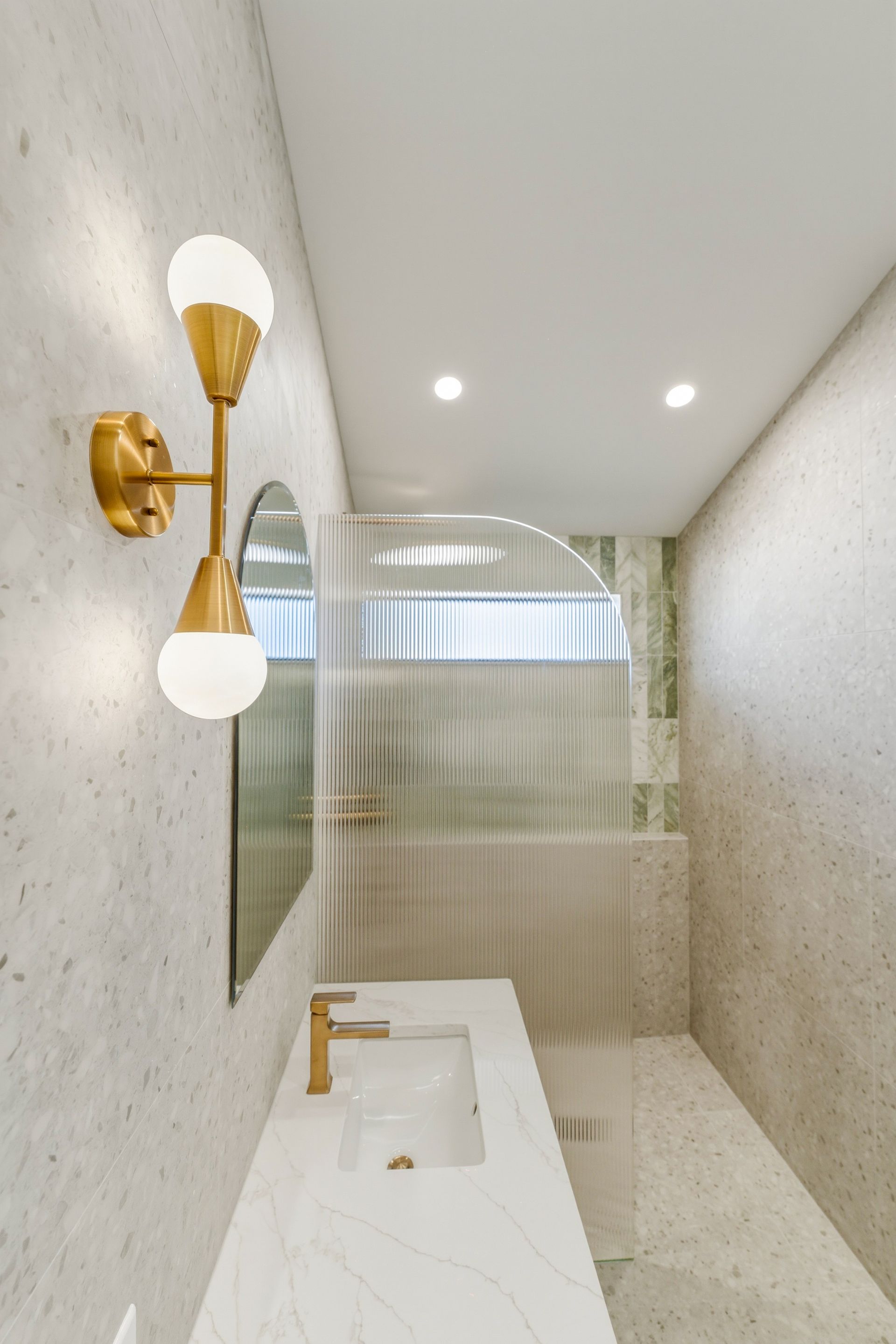 Narrow beige bathroom with white sink vanity, gold sconces, and frosted glass shower enclosure
