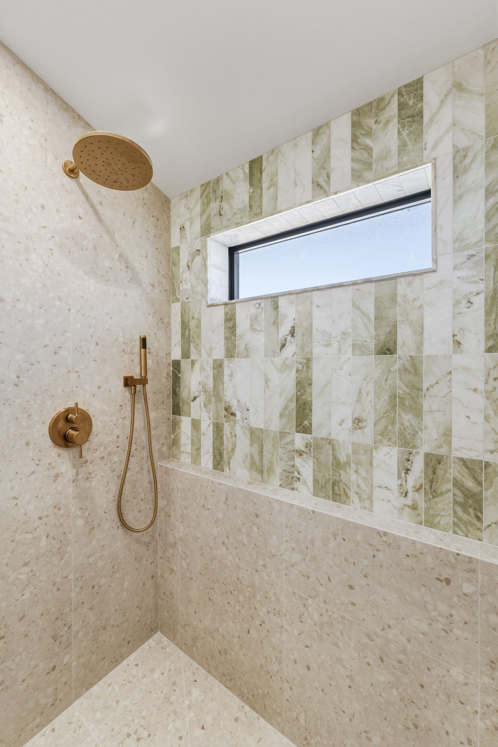 Modern shower with beige stone tile, gold fixtures, and a small horizontal window.