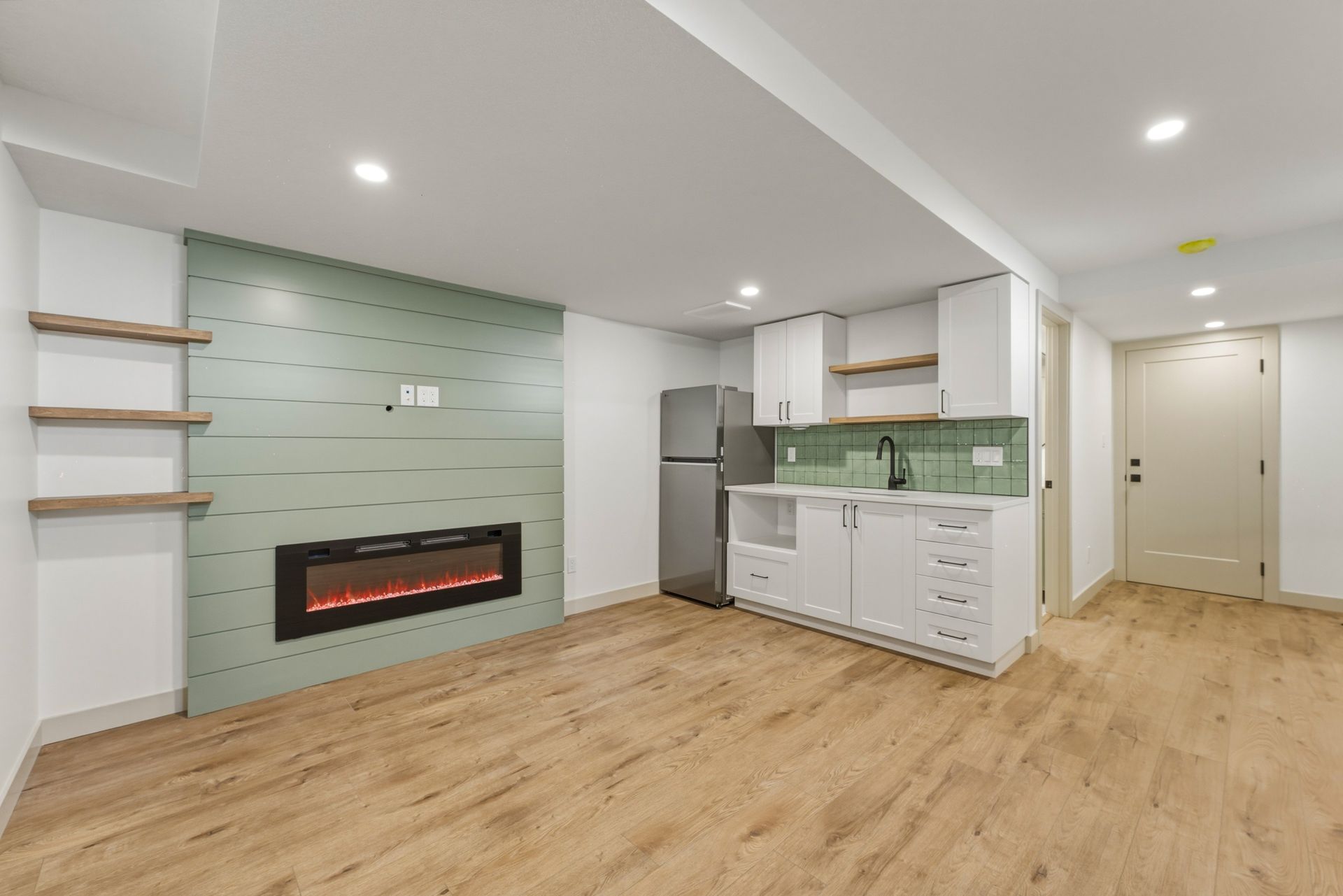 Modern open-plan living room with light wood floors, white cabinets, green tile accent wall, and electric fireplace