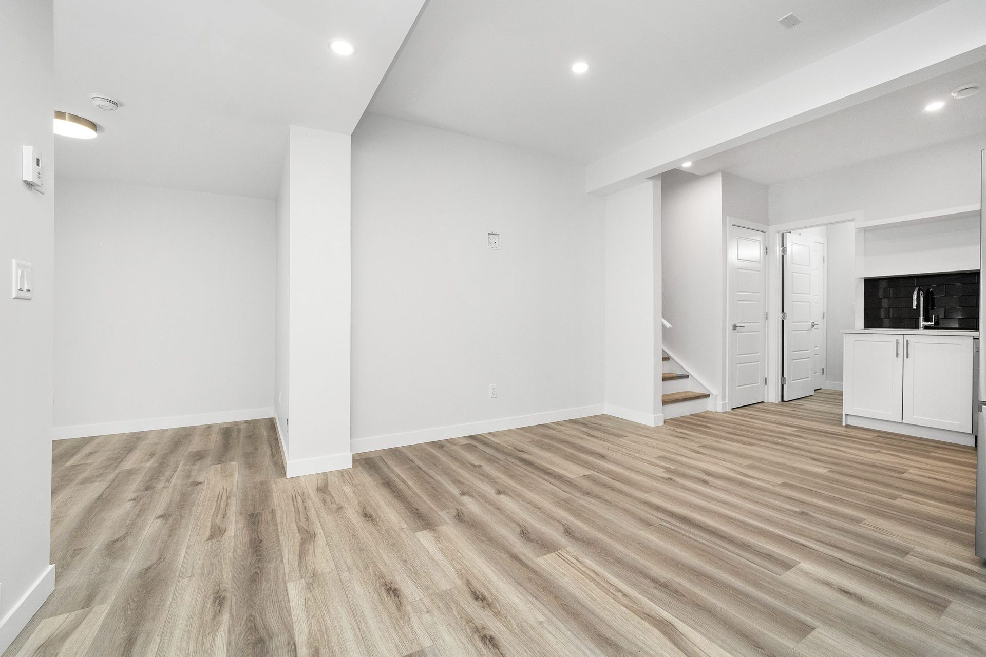 Empty modern open-plan room with light wood flooring, white walls, and a compact kitchen area