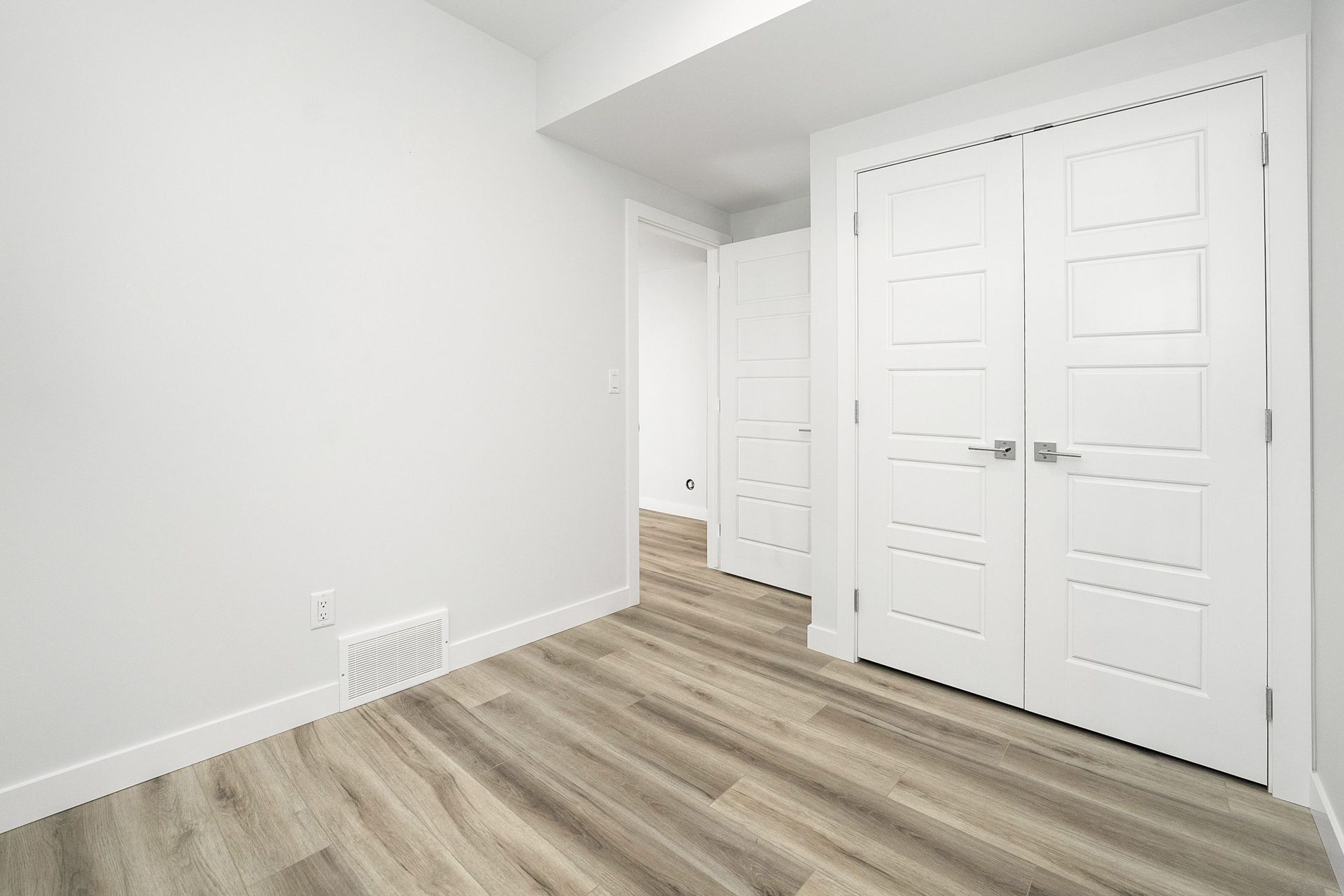 Empty white bedroom with wood floors and double closet doors, plus an open doorway.