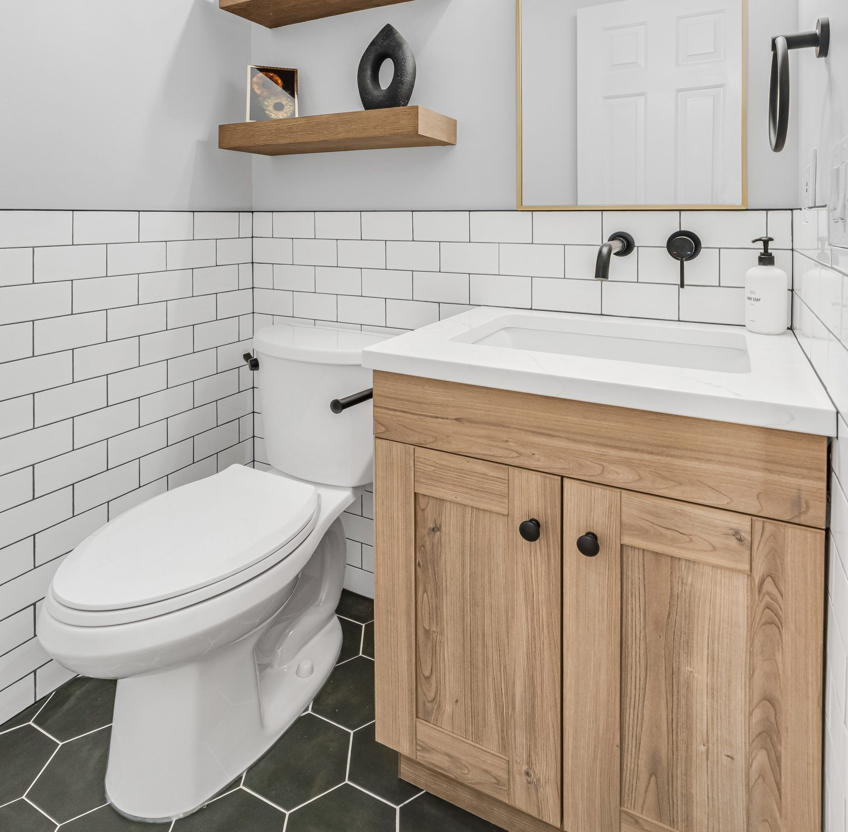 Small modern bathroom with white subway tile, wooden vanity, toilet, and black hexagon floor tiles