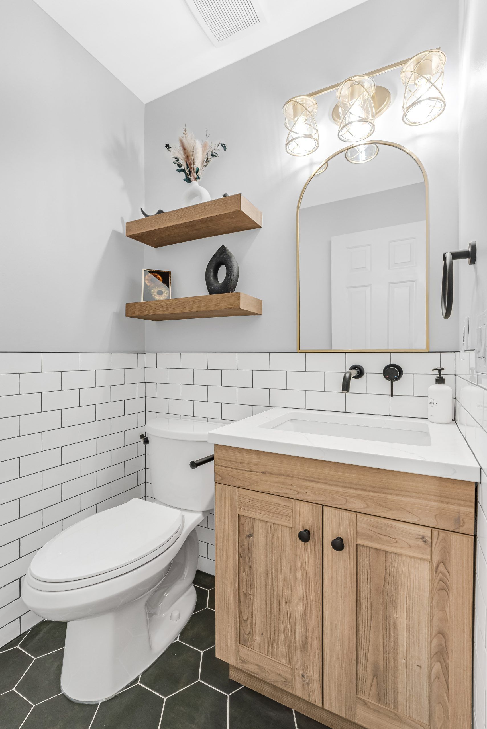 Modern bathroom with white tile, wood vanity, toilet, arched mirror, and floating shelves.