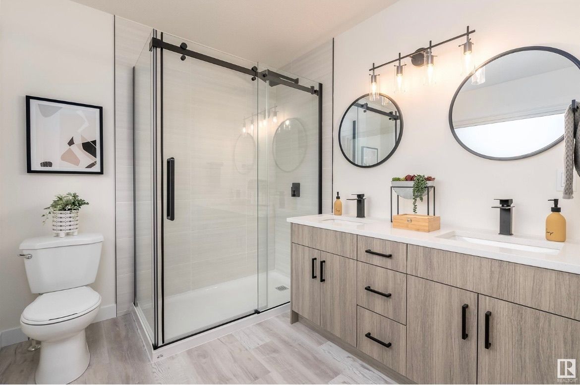 Modern bathroom with glass shower, white vanity, round mirrors, and toilet in a bright neutral space