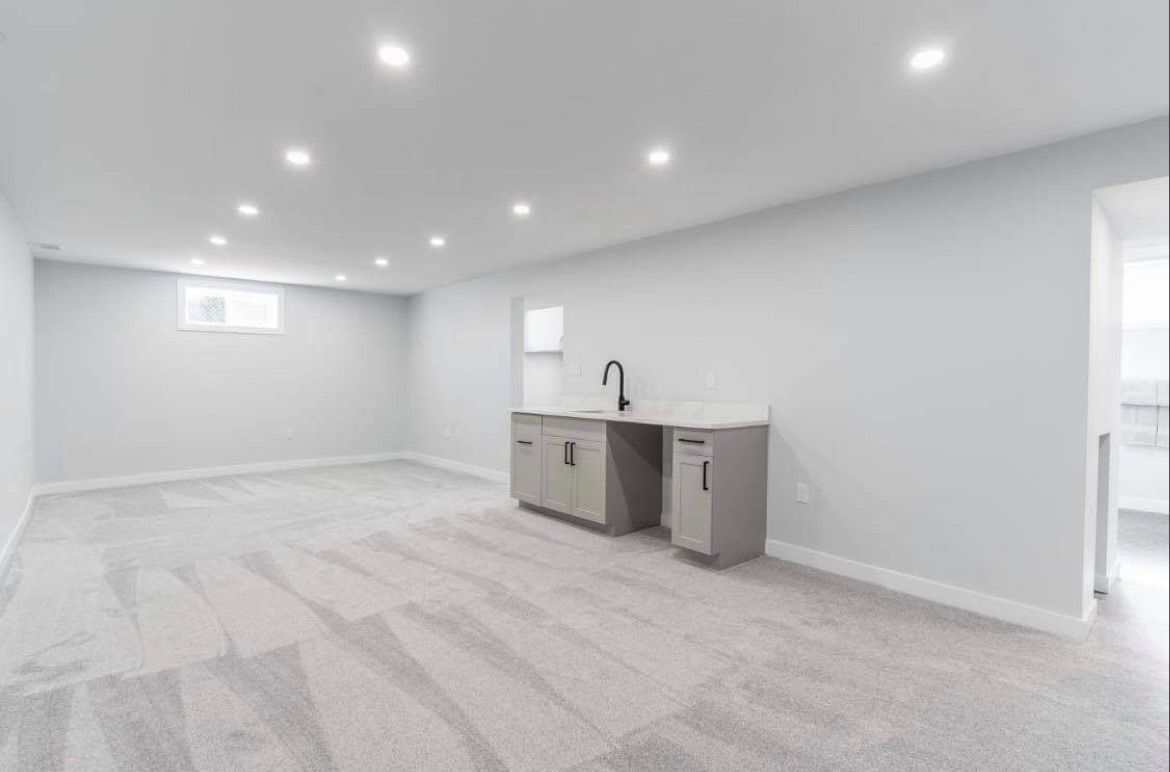 Bright empty basement room with white carpet, recessed lights, and a small gray kitchenette counter