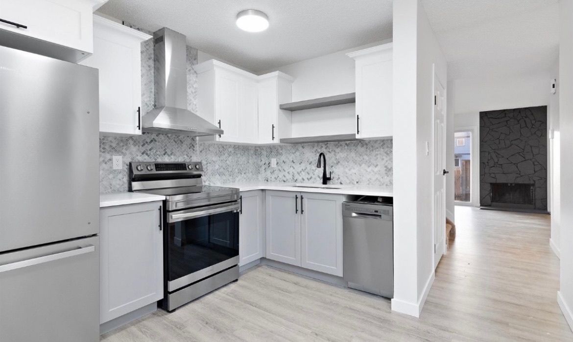 Modern white kitchen with stainless steel appliances, light wood floors, and a gray tile backsplash