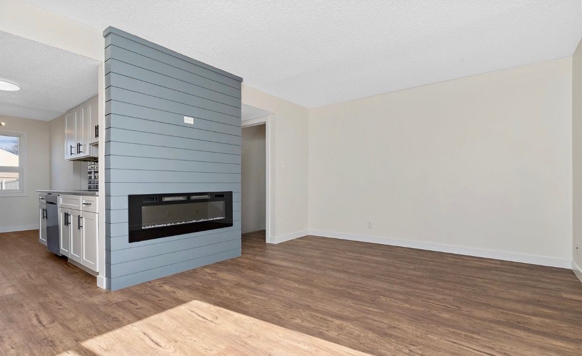 Empty living room with blue-tiled fireplace and wood floors, opening to a small kitchen.