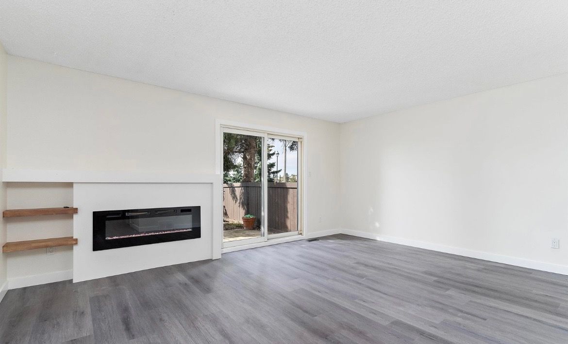 Empty modern living room with white walls, gray wood floors, fireplace, shelves, and sliding glass door to patio