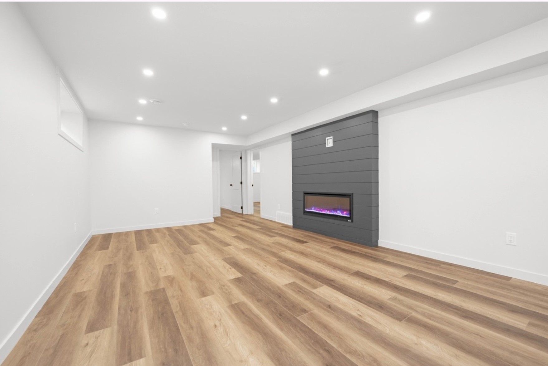 Spacious empty living room with hardwood floors, white walls, recessed lights, and a dark fireplace accent wall.